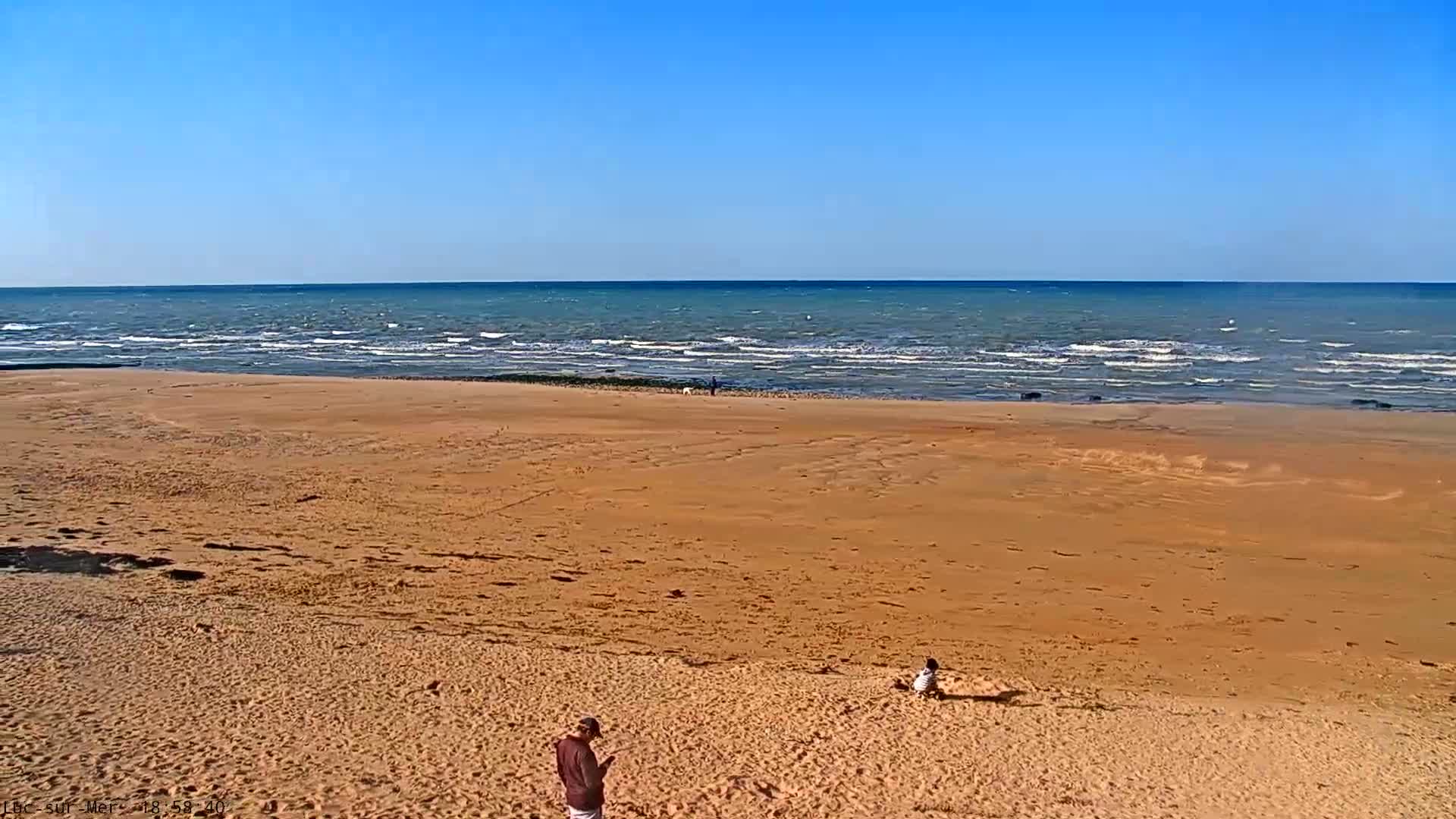 A wide sandy beach slopes down to a wavy ocean under a clear blue sky.