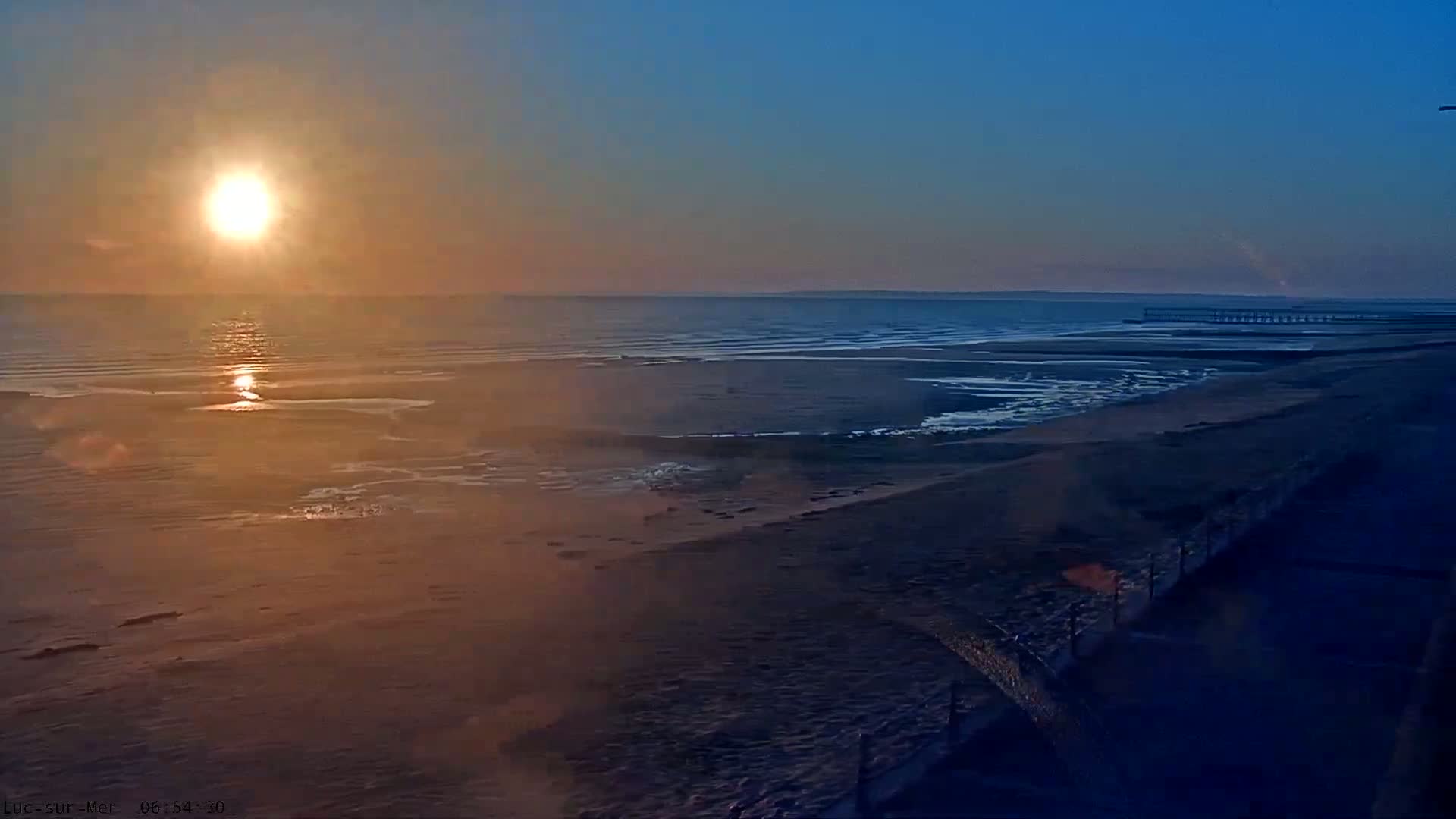 A sunrise casts a golden glow over a calm ocean beach, with a distant pier visible under a clear sky.