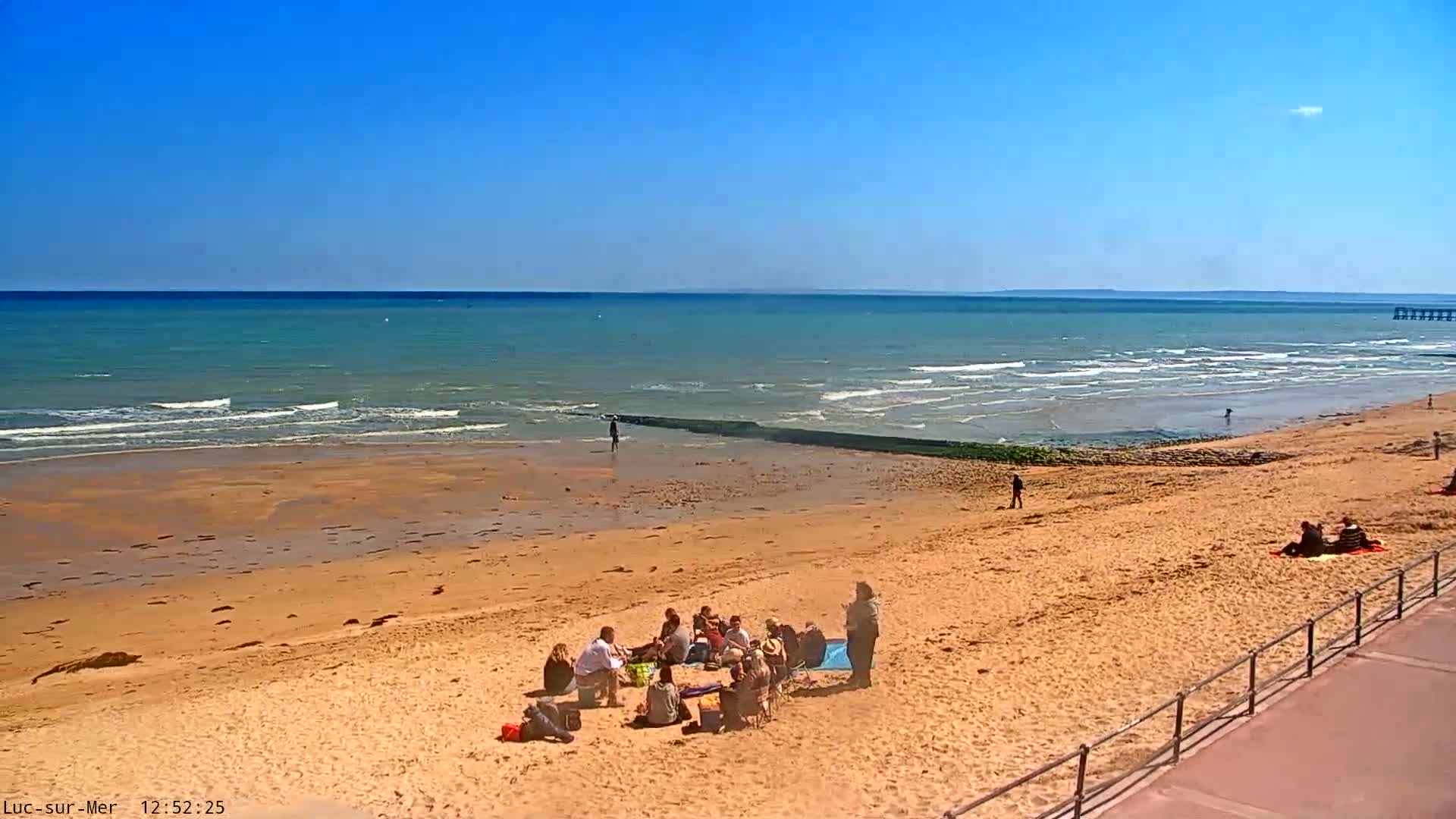 A sandy beach with gentle waves under a clear blue sky, showing a group of people sitting together on the sand.