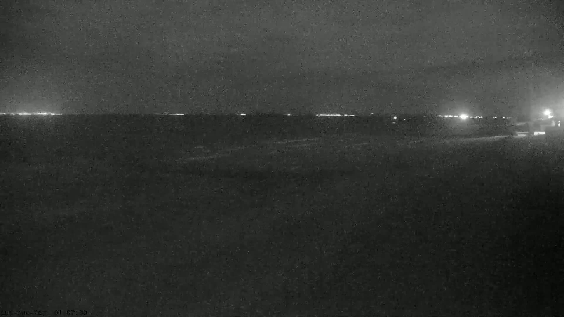 A nighttime, low-light view shows a dark body of water with a distant line of lights on the horizon.