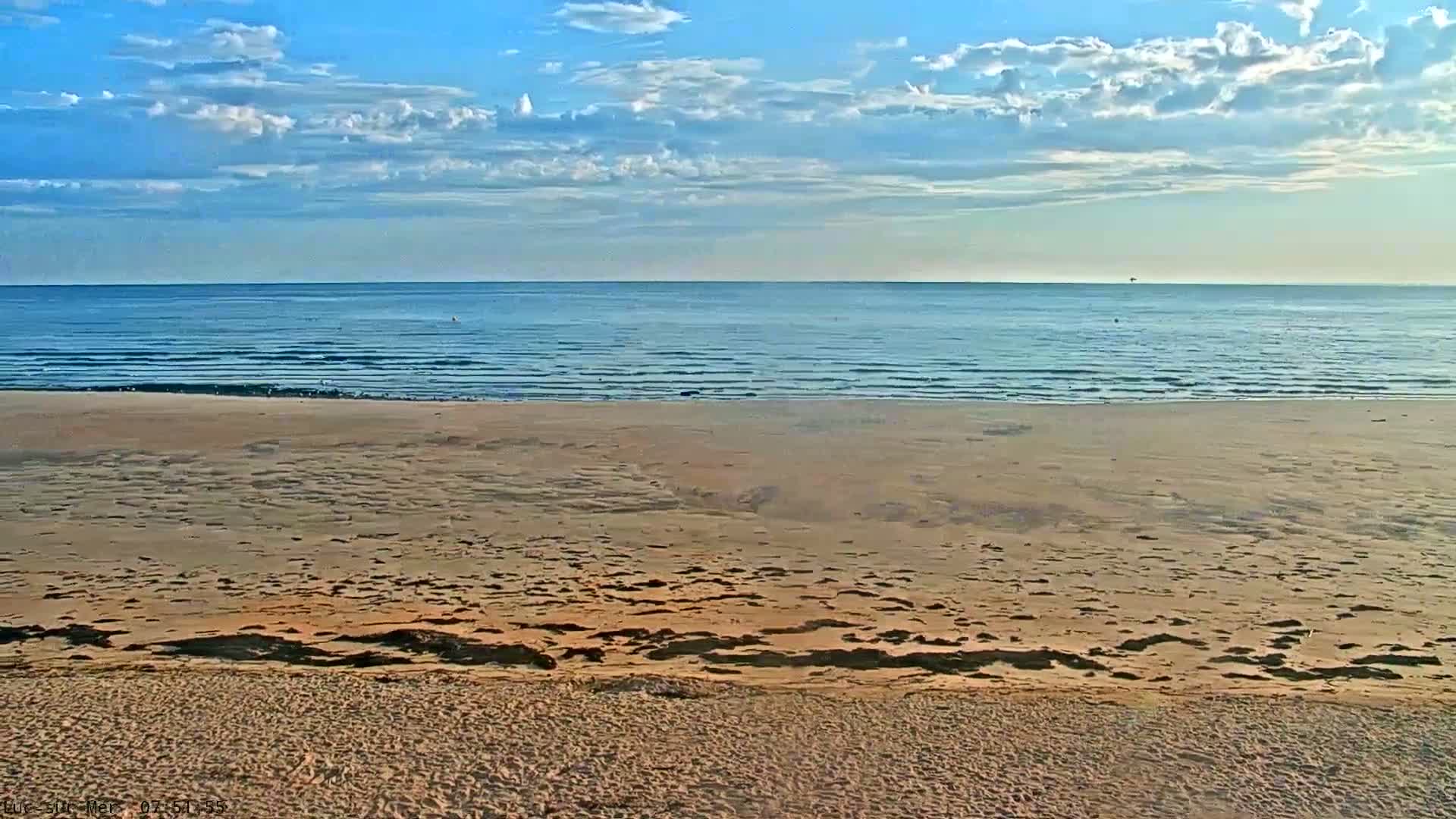 A sandy beach stretches to a calm ocean under a partly cloudy blue sky.