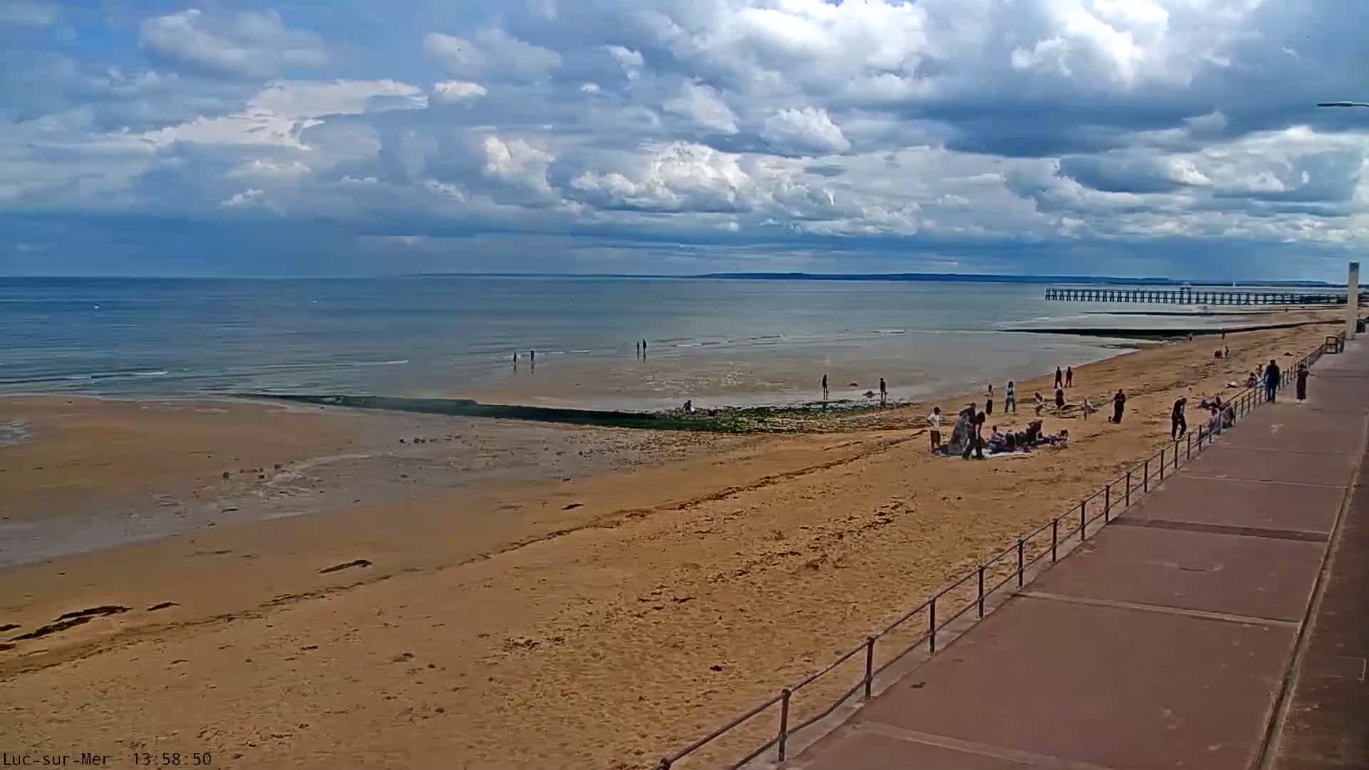 A sandy beach with scattered people, a pier in the distance, and a partly cloudy sky.