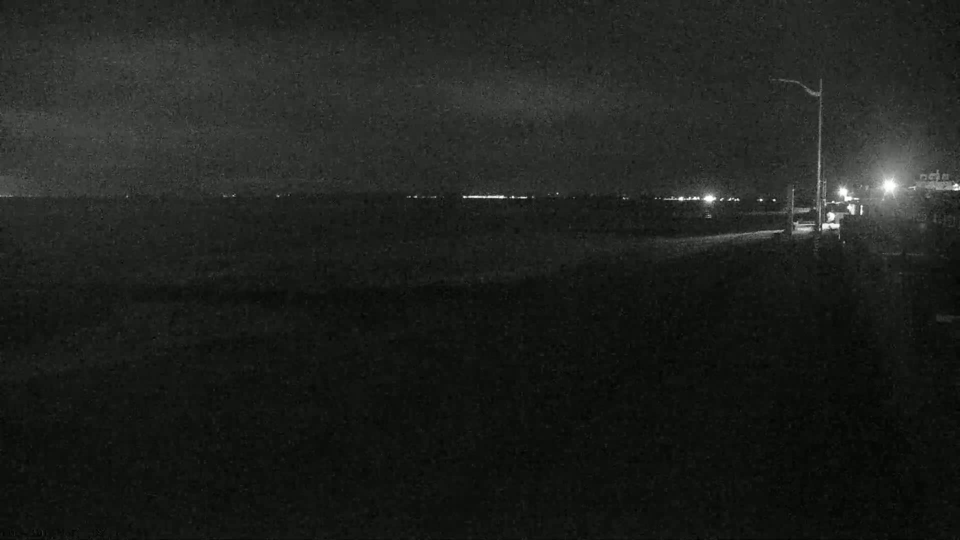 A nighttime view of a dark beach with a faint line of lights in the distance under a cloudy sky.