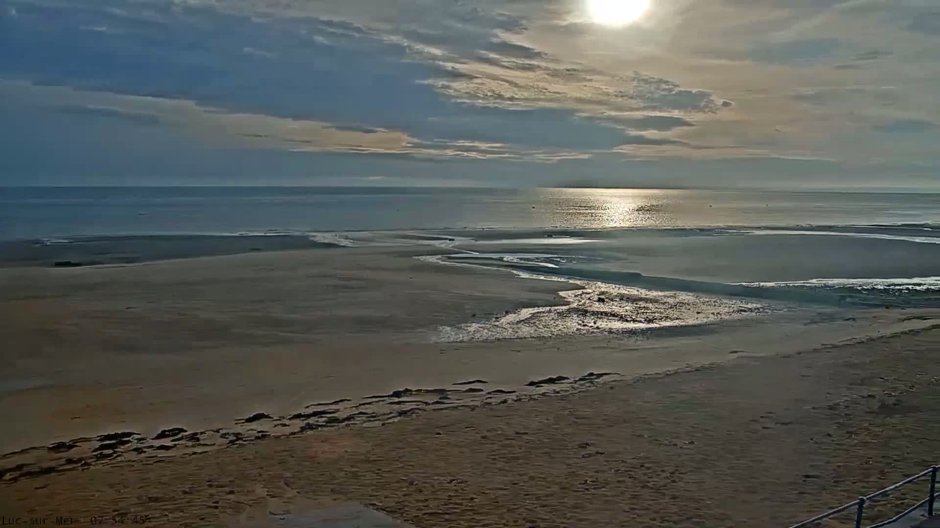 A sandy beach with tidal streams leading to a calm sea under a partly cloudy sky with the sun shining brightly.