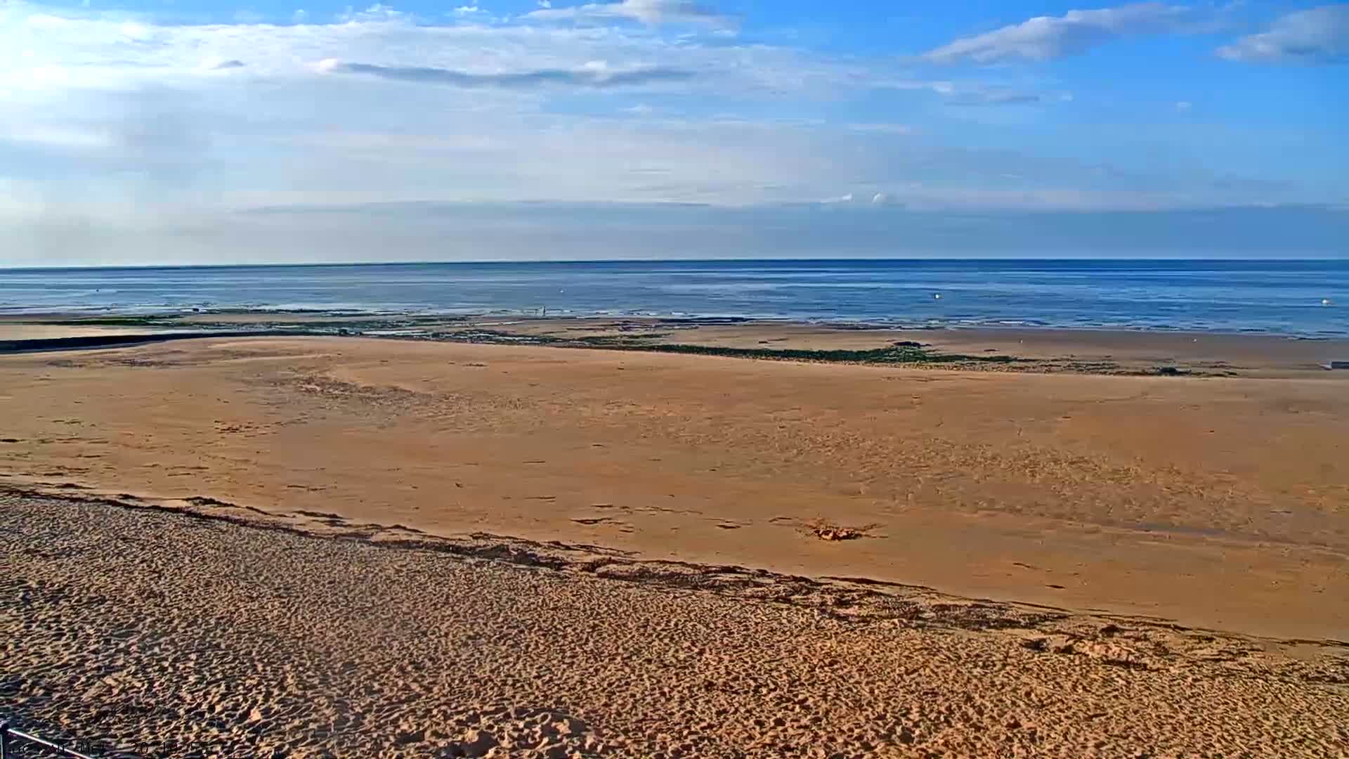 A wide expanse of sandy beach stretches towards a calm ocean under a mostly clear, bright sky.