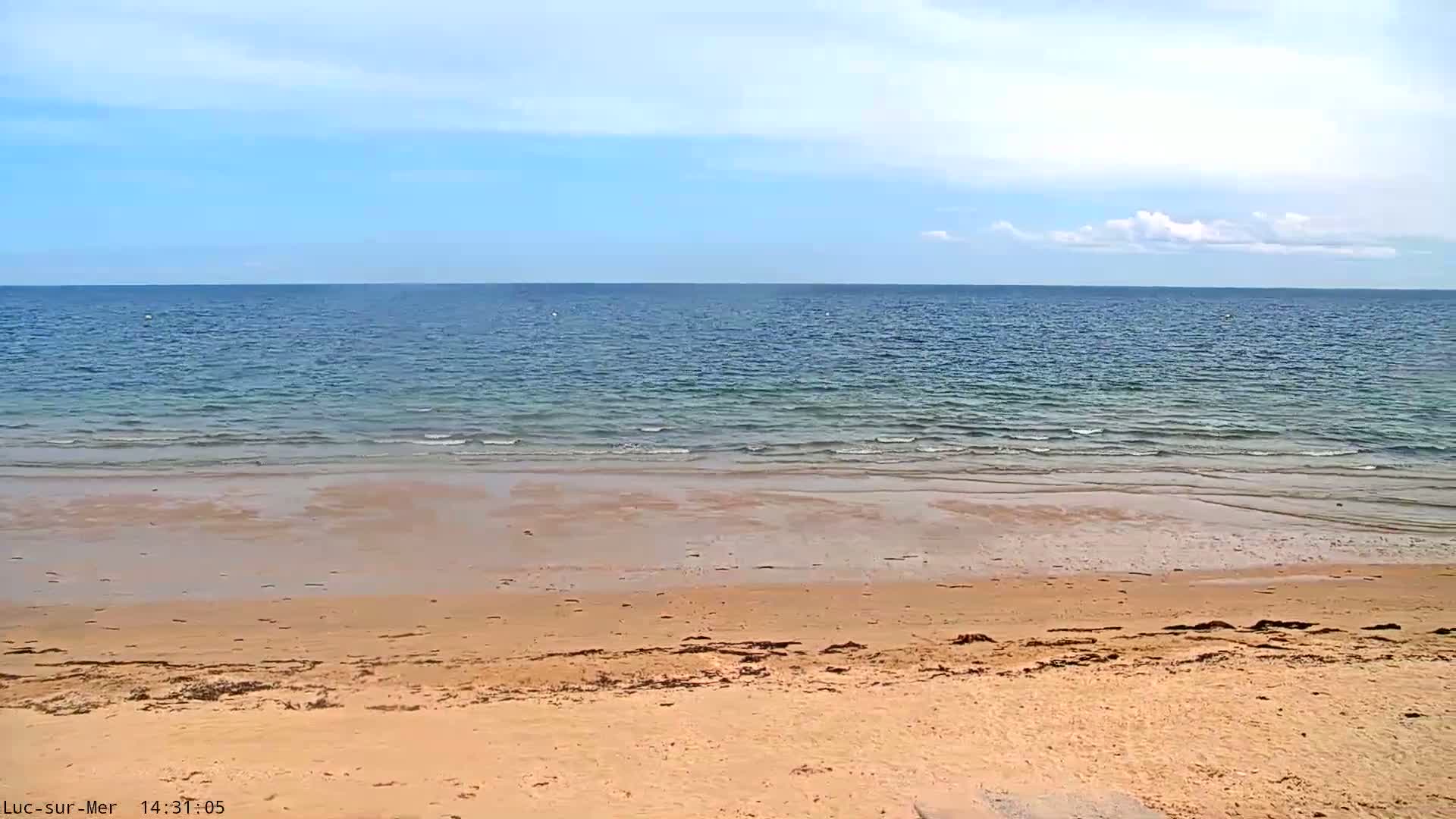 A sandy beach slopes down to a calm, blue ocean under a mostly clear sky with a few scattered clouds.