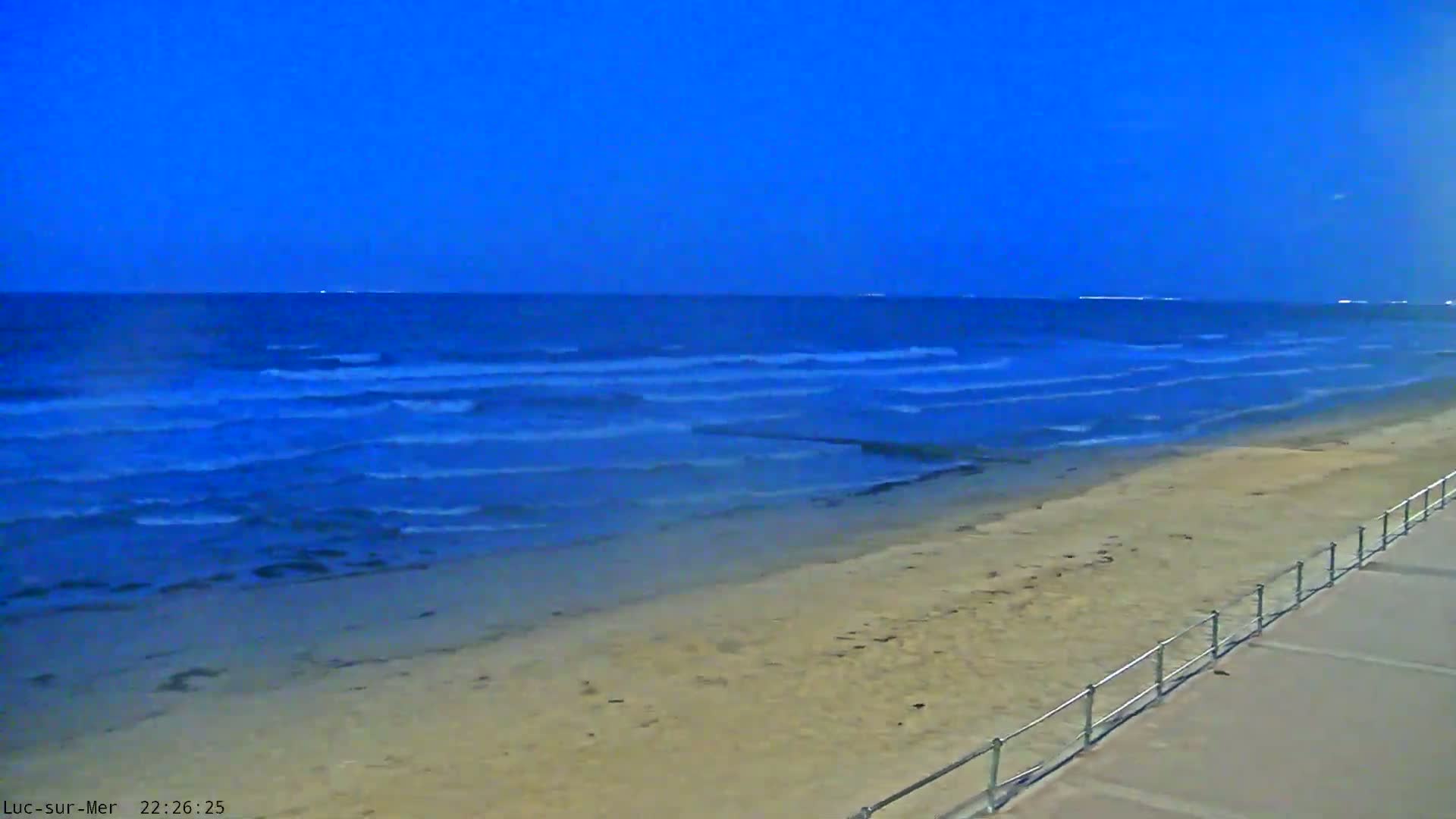 Under a dark blue sky, gentle waves lap a sandy beach next to a walkway with a railing.