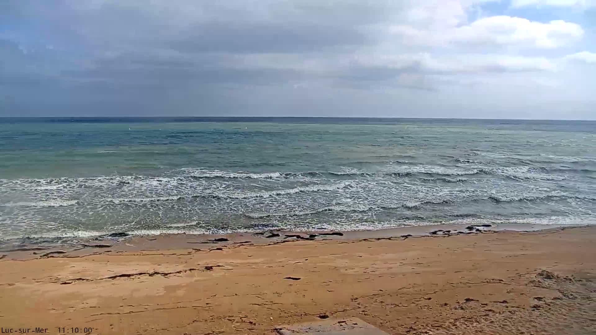 A sandy beach slopes down to a sea with small waves under a mostly cloudy sky.