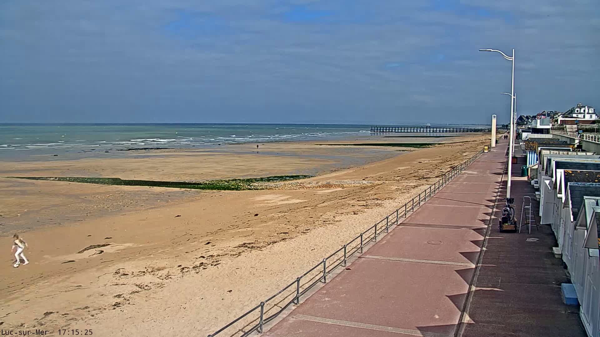 A sandy beach with a boardwalk and beach huts stretches alongside a calm ocean under a partly cloudy sky.