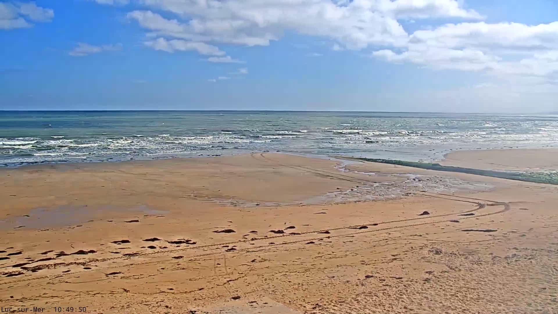 A sandy beach with tire tracks leading to a calm ocean under a partly cloudy sky.