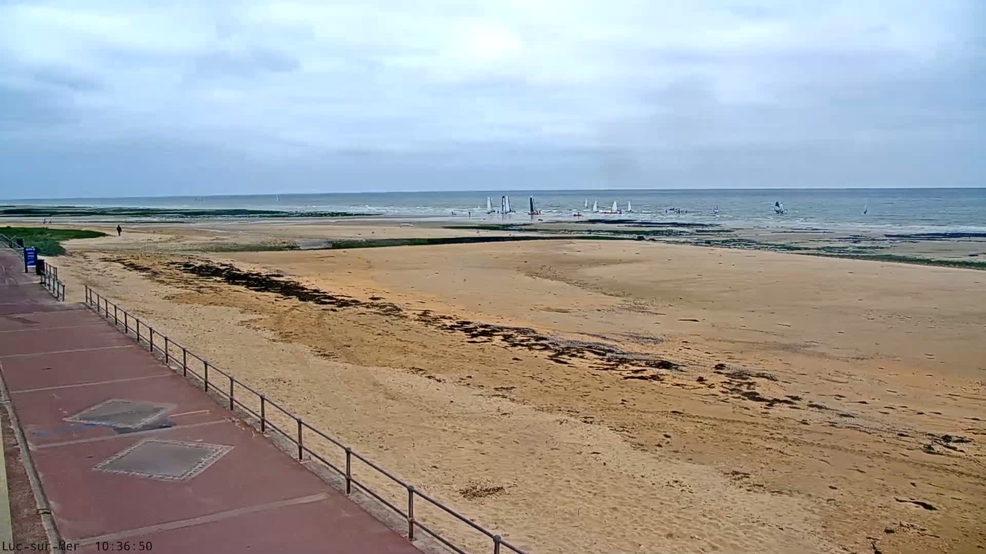 A wide sandy beach with a paved walkway and railing stretches toward a calm ocean where several sailboats are visible under an overcast sky.