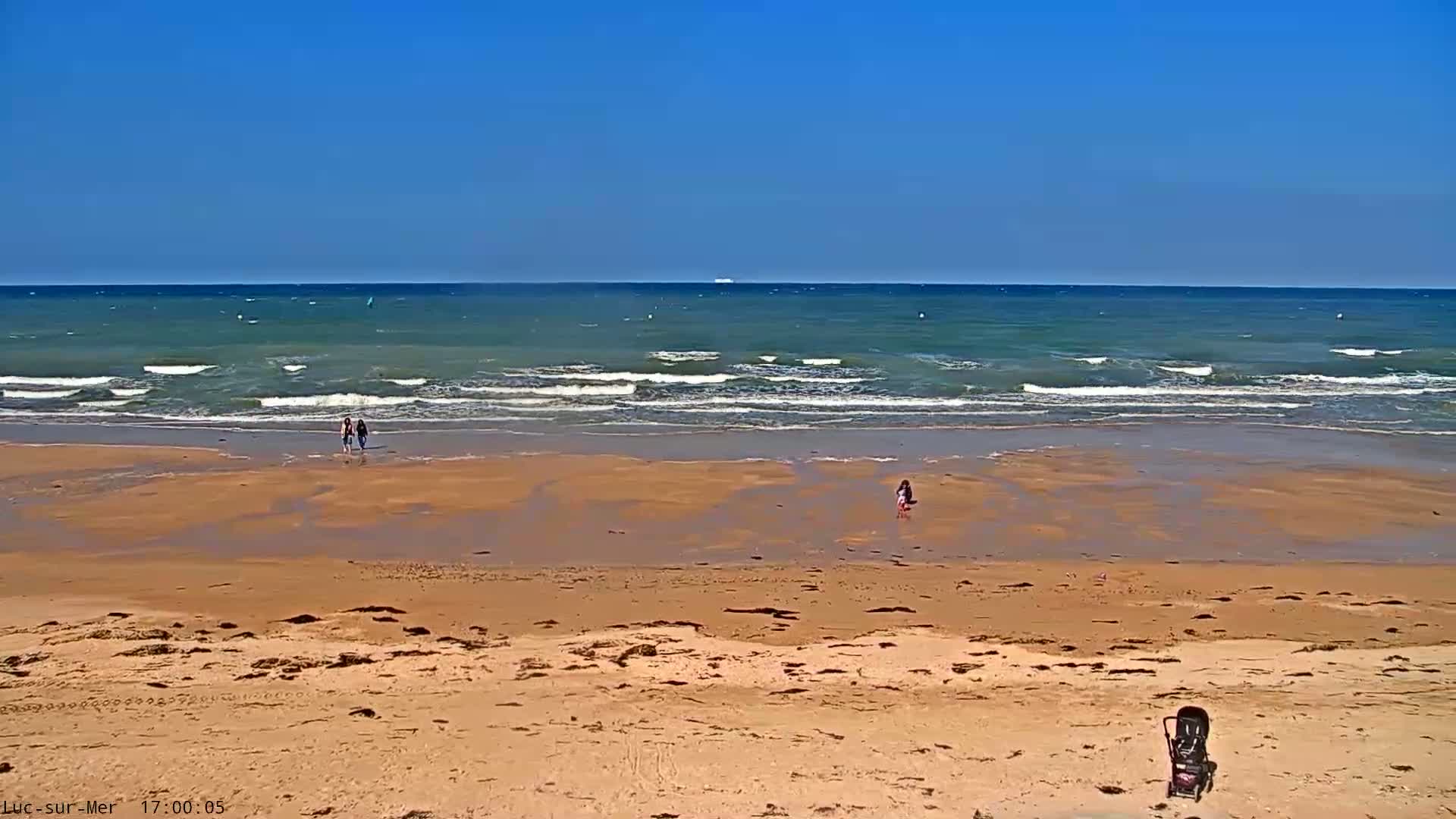 A sandy beach with a few people walking near the water's edge under a clear blue sky, with small waves rolling onto the shore.