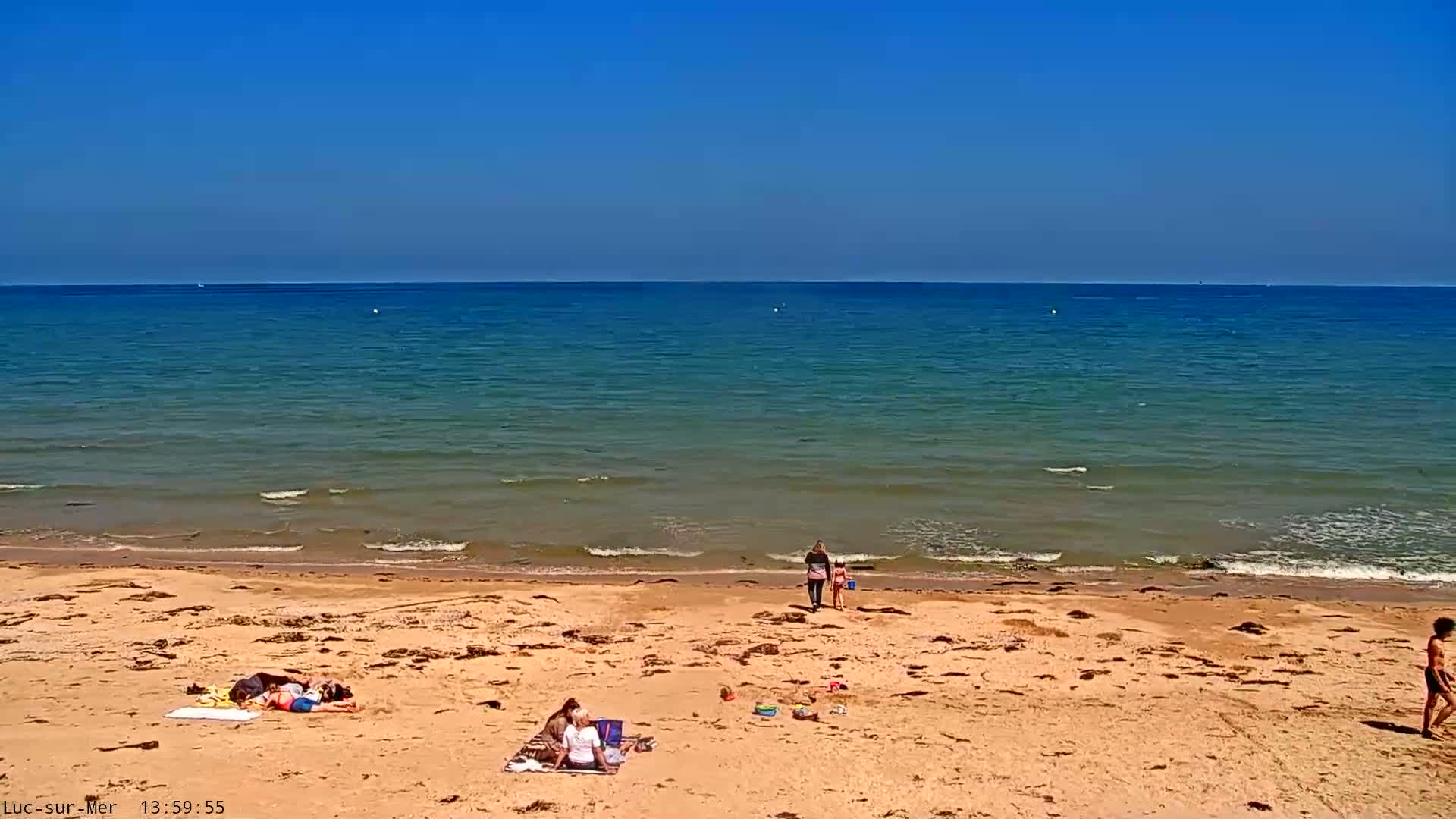 A sandy beach with several people relaxing and playing near calm, blue water under a clear blue sky.