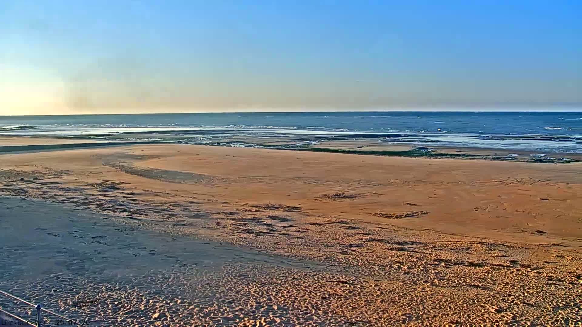 A wide sandy beach stretches to a calm ocean under a clear blue sky.