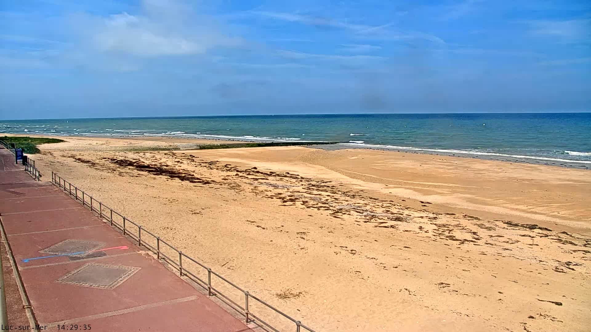 A sandy beach with a reddish-brown paved walkway and railing stretches toward a calm, blue ocean under a mostly sunny sky.