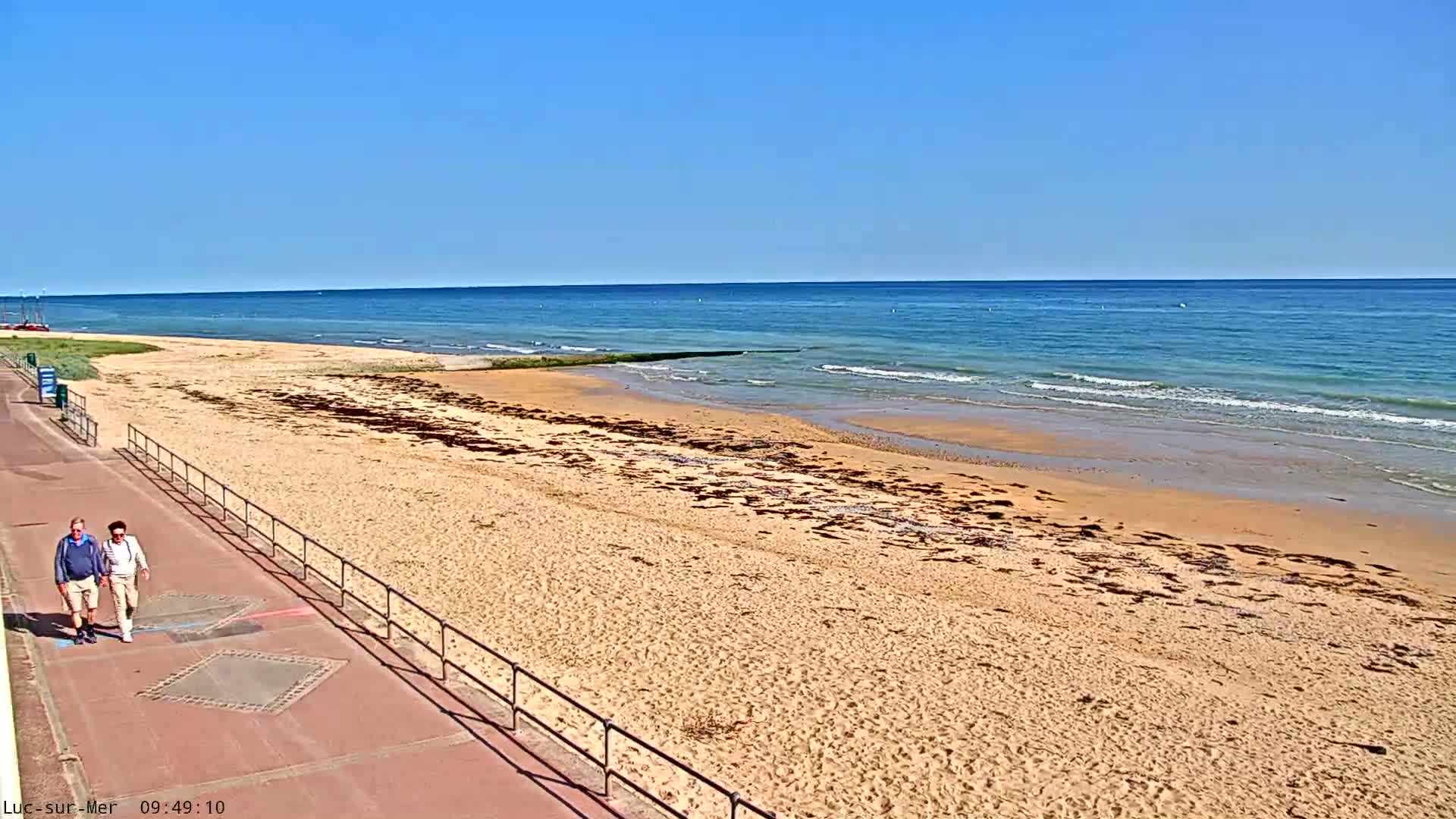 Two people walk along a paved beachfront path next to a sandy beach and calm, blue ocean under a clear sky.
