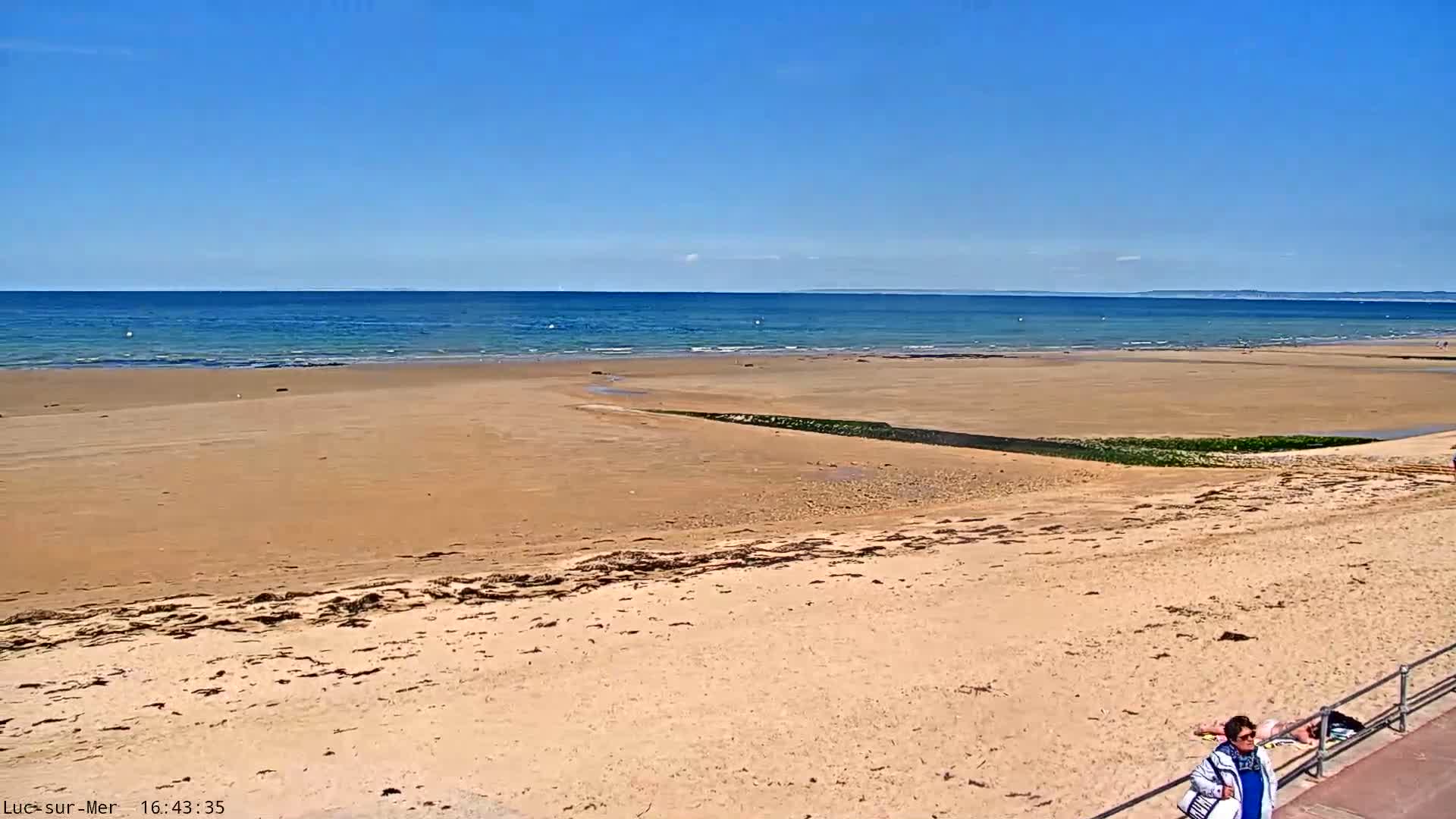 A wide sandy beach stretches to a calm, blue ocean under a clear blue sky, with a single person visible near a railing in the bottom right corner.