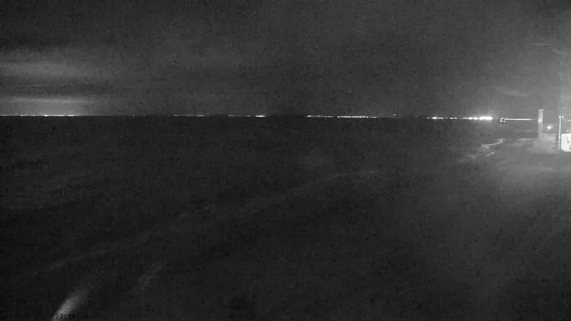A nighttime, low-light view of a dark body of water with a faint, distant city skyline visible on the horizon under a cloudy sky.