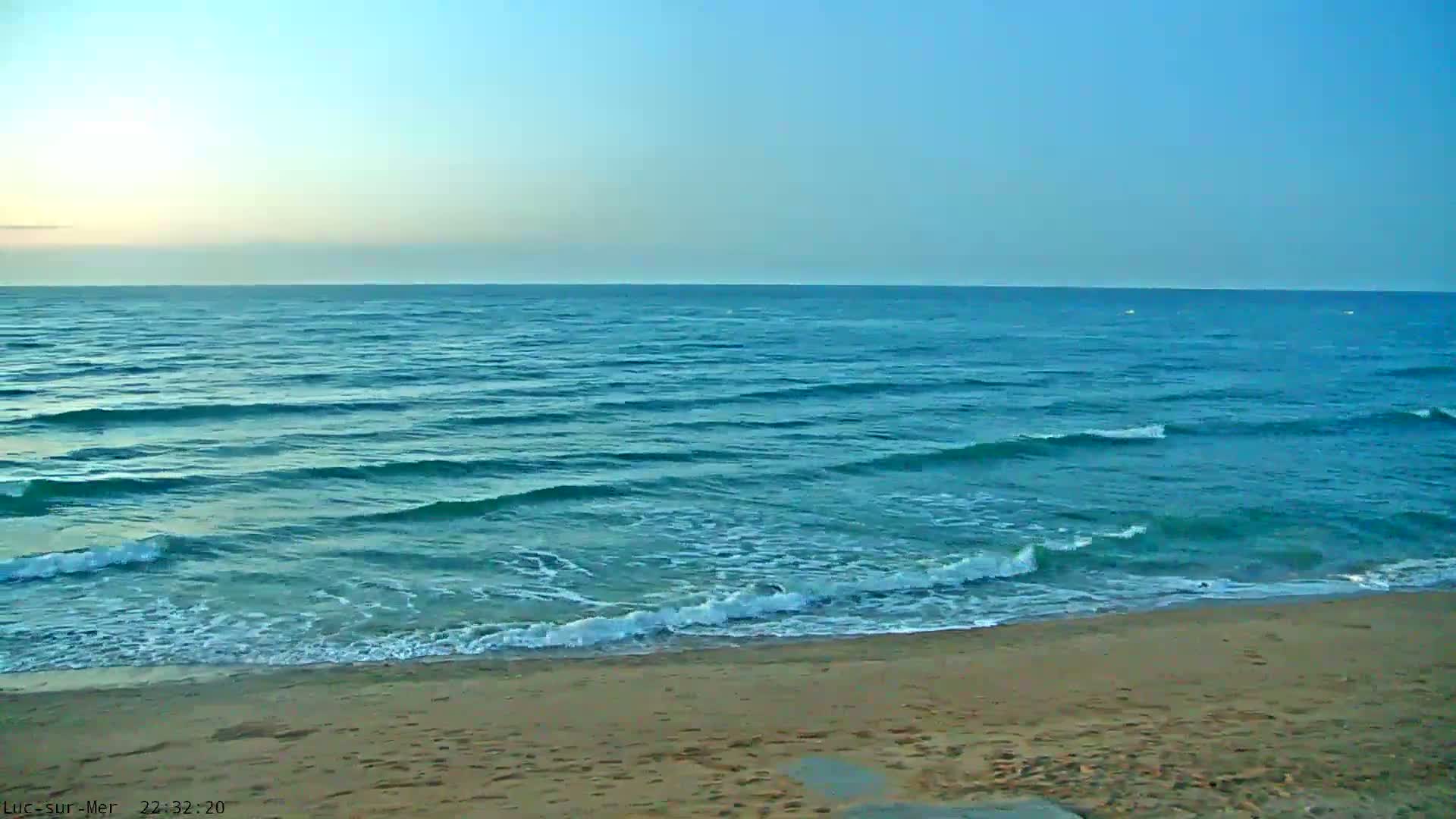 A sandy beach meets a calm, teal ocean under a clear, light-blue sky.