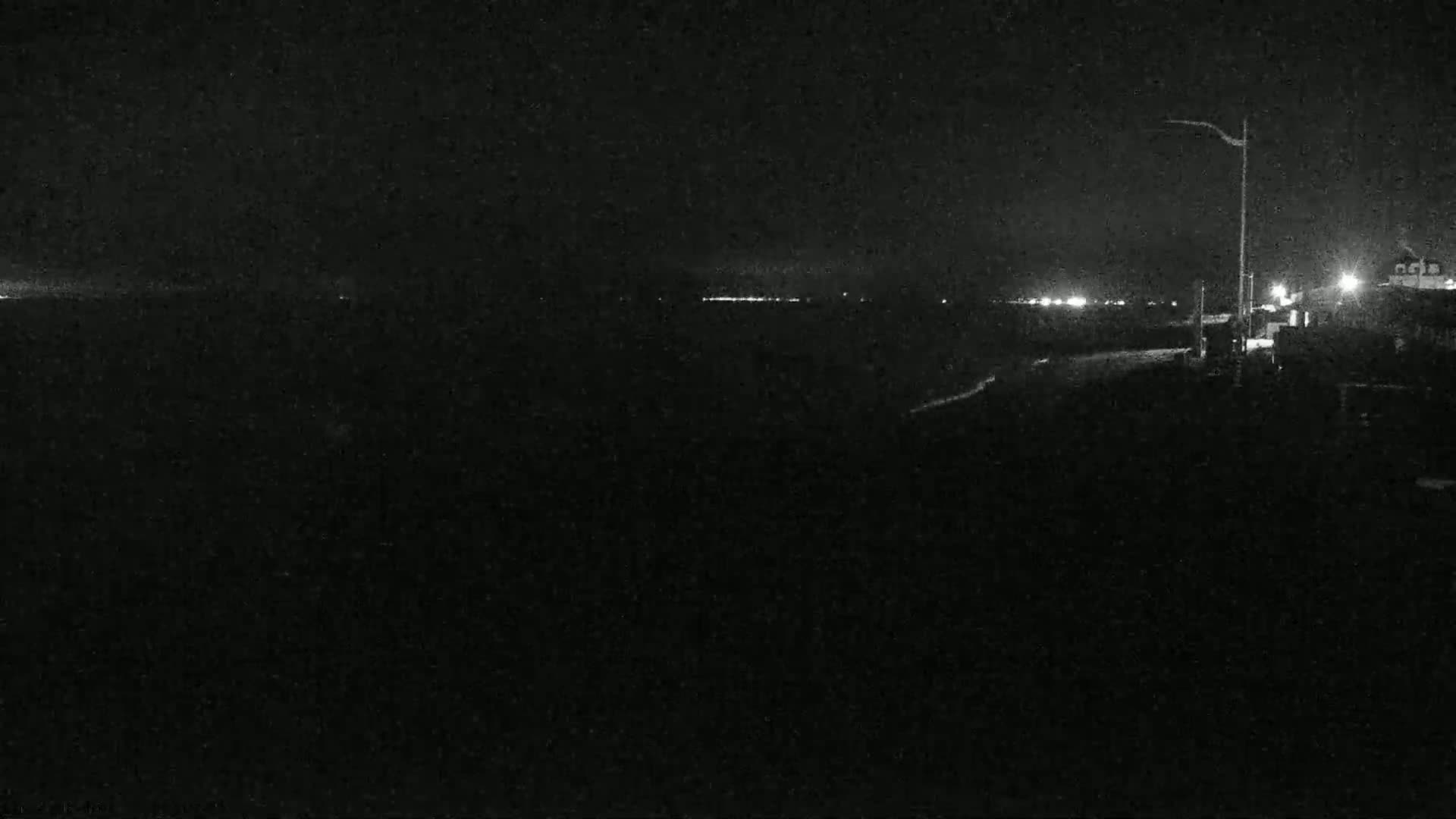 A nighttime view of a dark shoreline with distant lights and a single lamppost.
