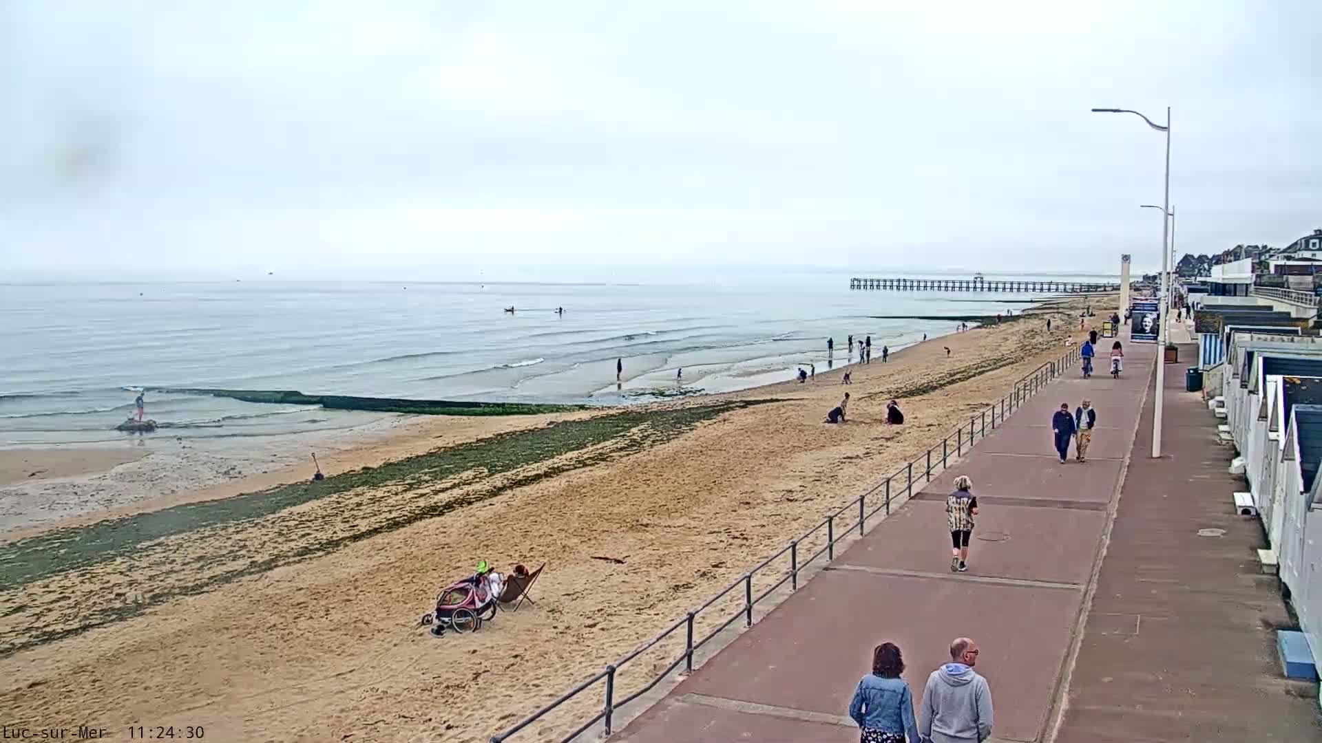 A sandy beach with a few people on it and a paved walkway next to it stretches toward a pier on an overcast day.
