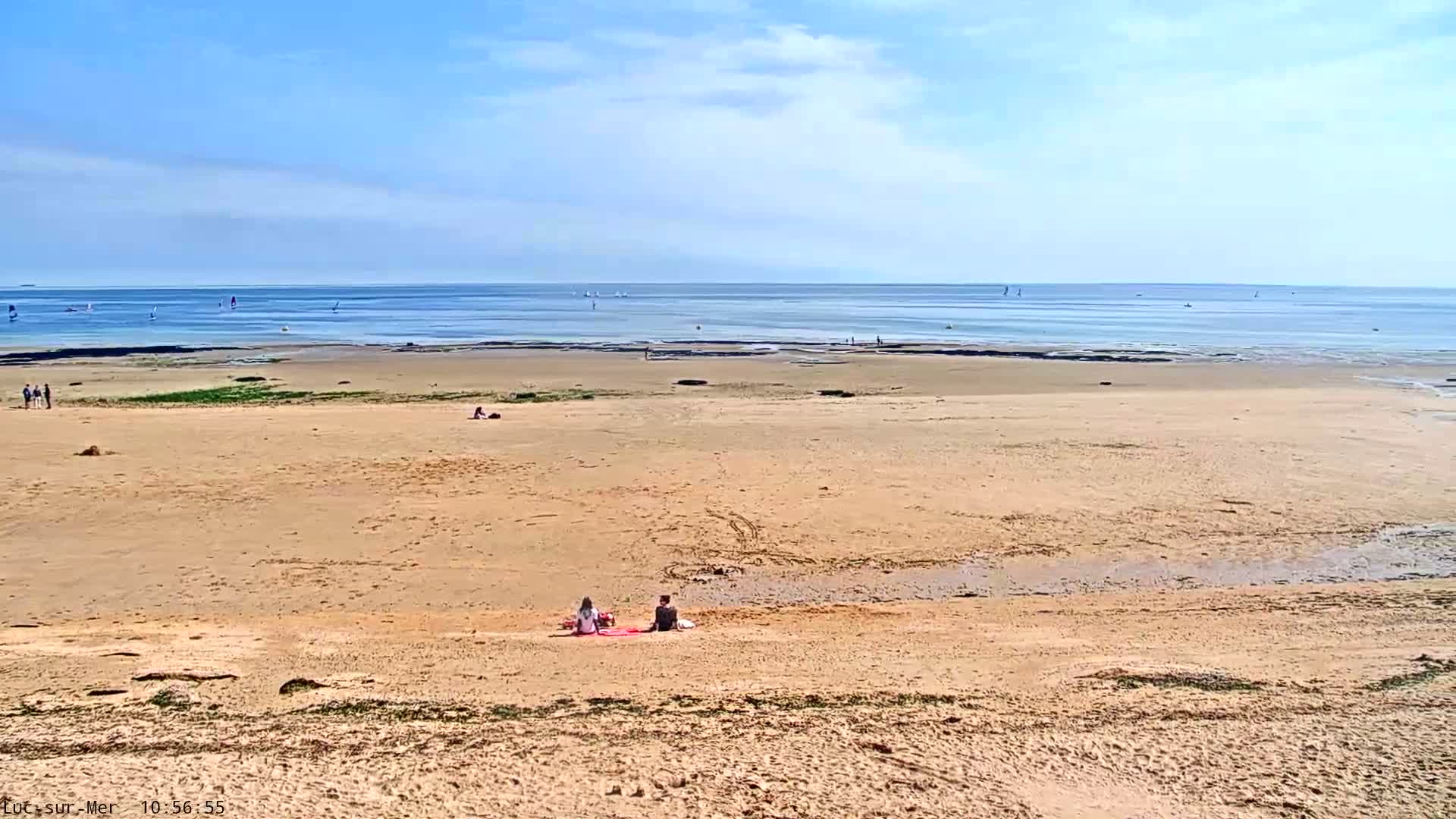 A sandy beach stretches to a calm, blue ocean under a clear sky, with a few people scattered on the sand and several sailboats visible on the water.