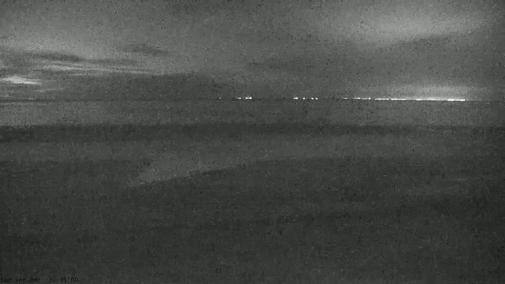 A dark, nighttime view shows a distant line of lights on the horizon over a dark body of water under a mostly clear sky.