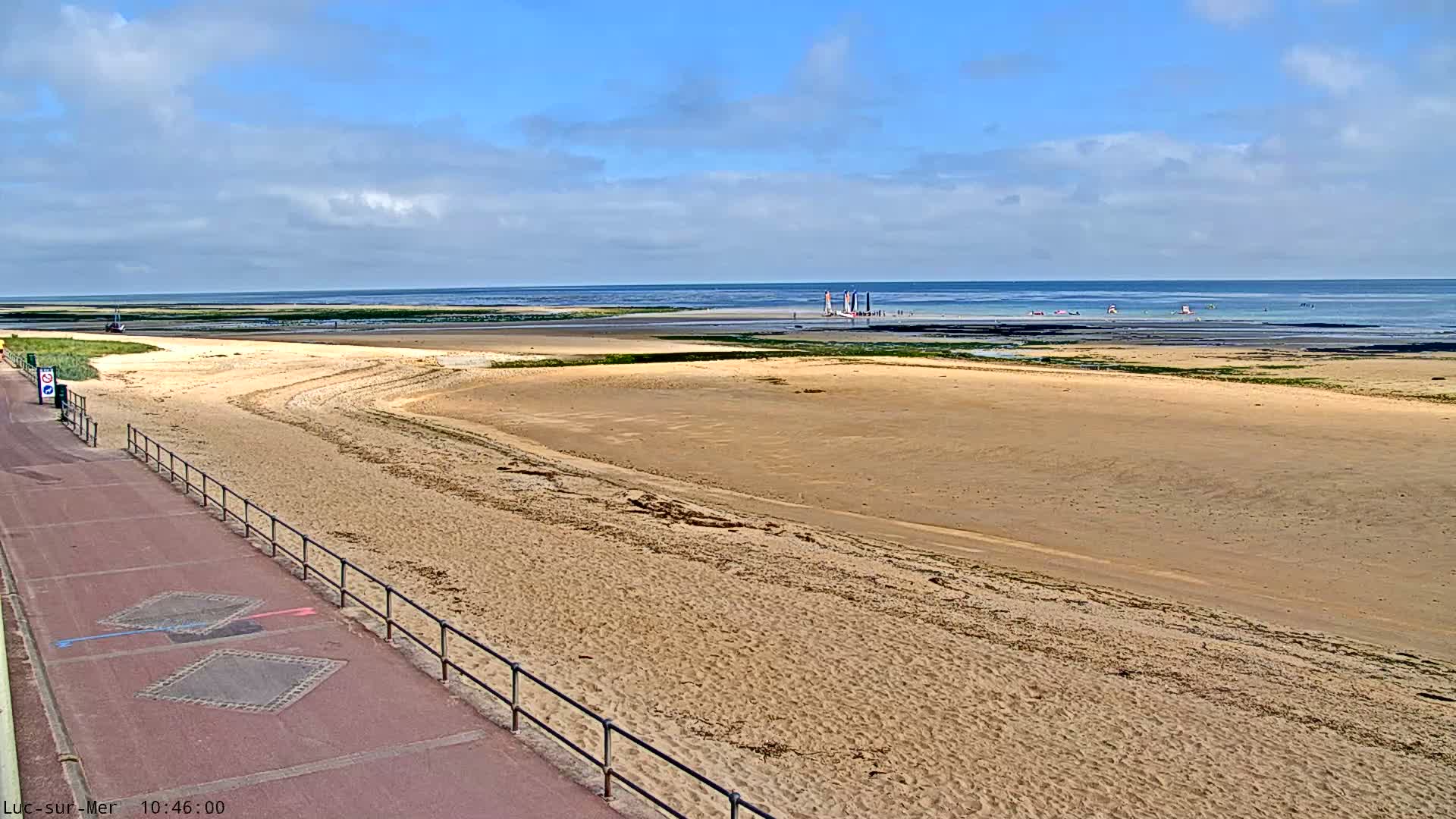A wide sandy beach stretches towards a calm ocean under a mostly sunny sky, with a few sailboats visible in the distance and a paved walkway along the beach's edge.