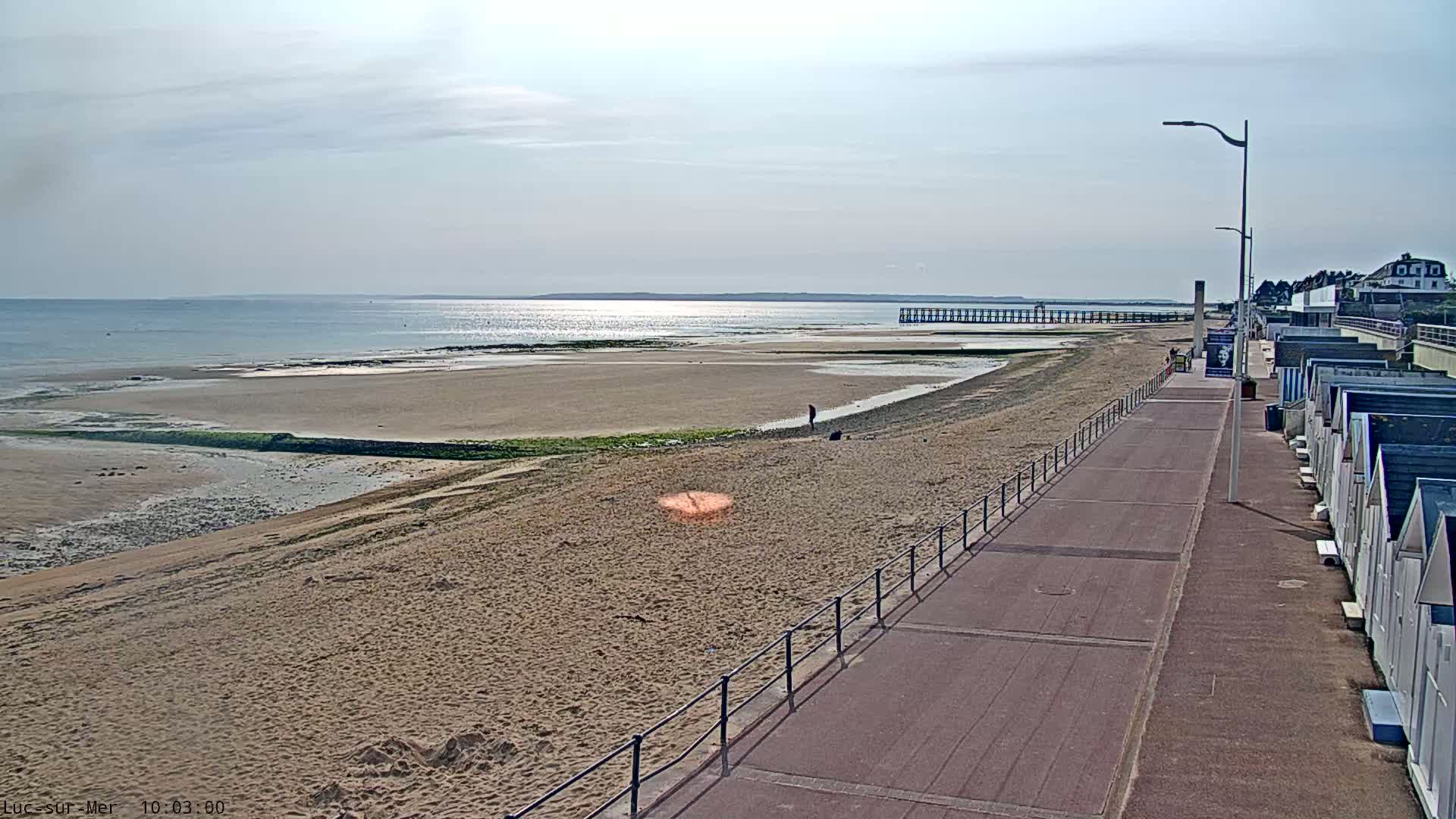 A wide sandy beach with a boardwalk and beach huts stretches alongside a calm sea under a partly cloudy sky.