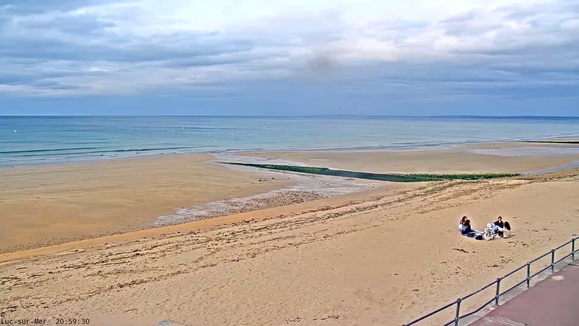 A sandy beach stretches to a calm ocean under a mostly cloudy sky, with a small group of people sitting together near the water's edge.