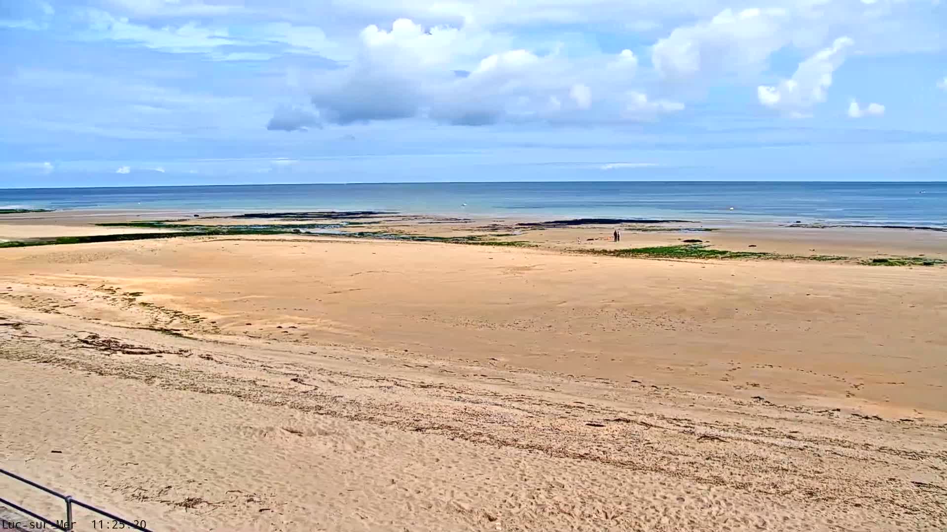 A mostly sunny day reveals a wide expanse of sandy beach sloping down to a calm ocean under a partly cloudy sky.
