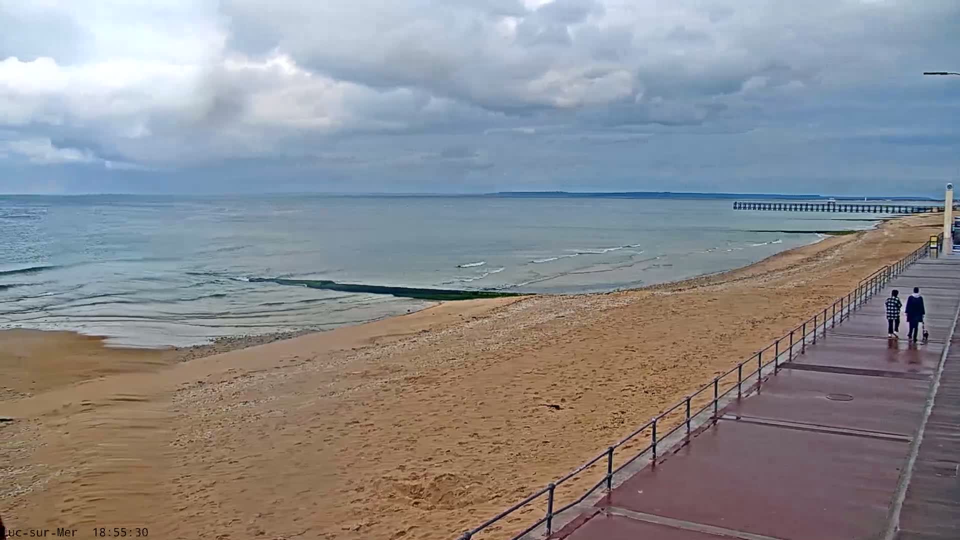 Two people walk a boardwalk alongside a sandy beach and calm ocean under a cloudy sky.