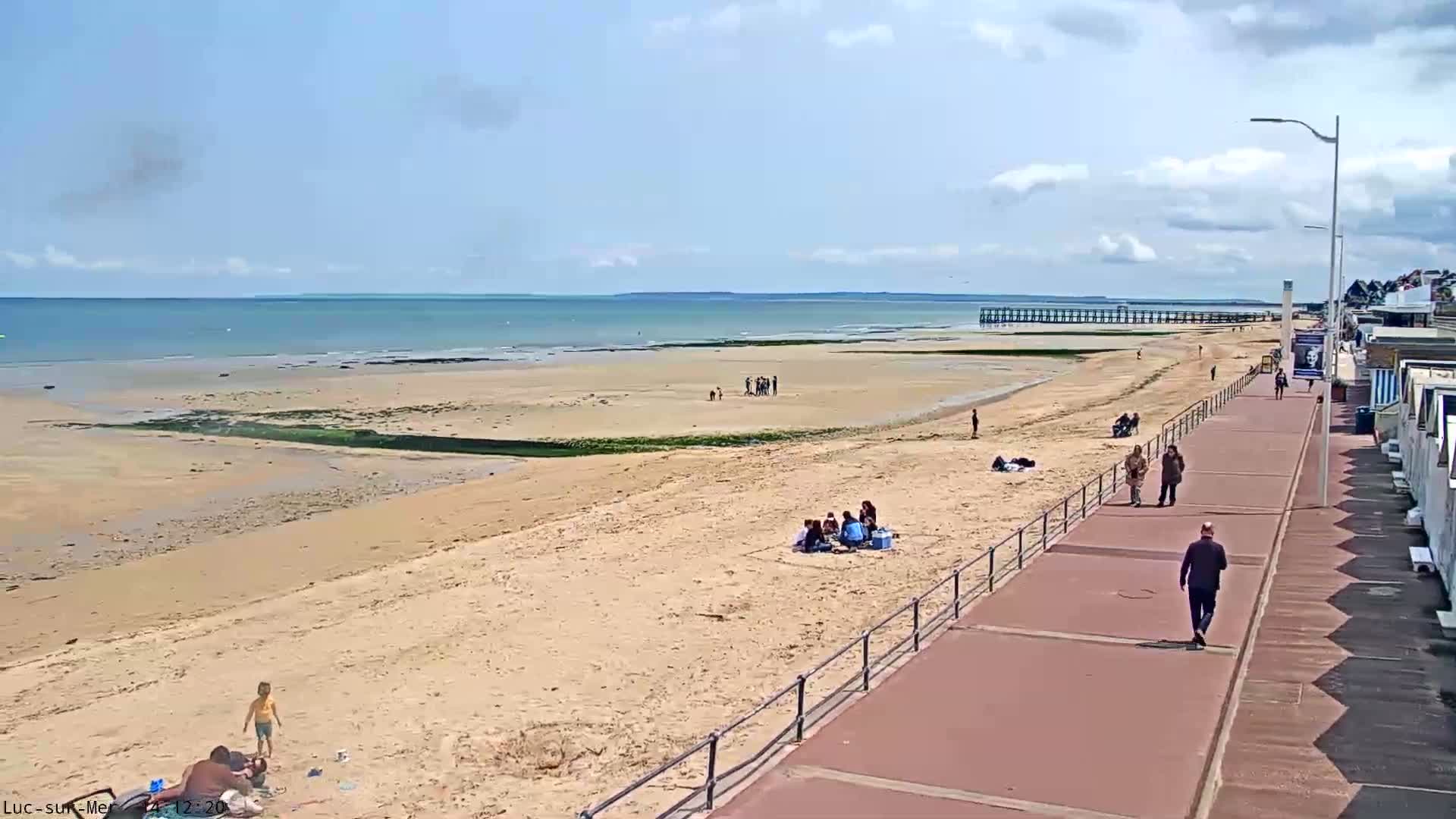 A sandy beach with people relaxing and walking along a boardwalk, under a mostly sunny sky with some clouds.