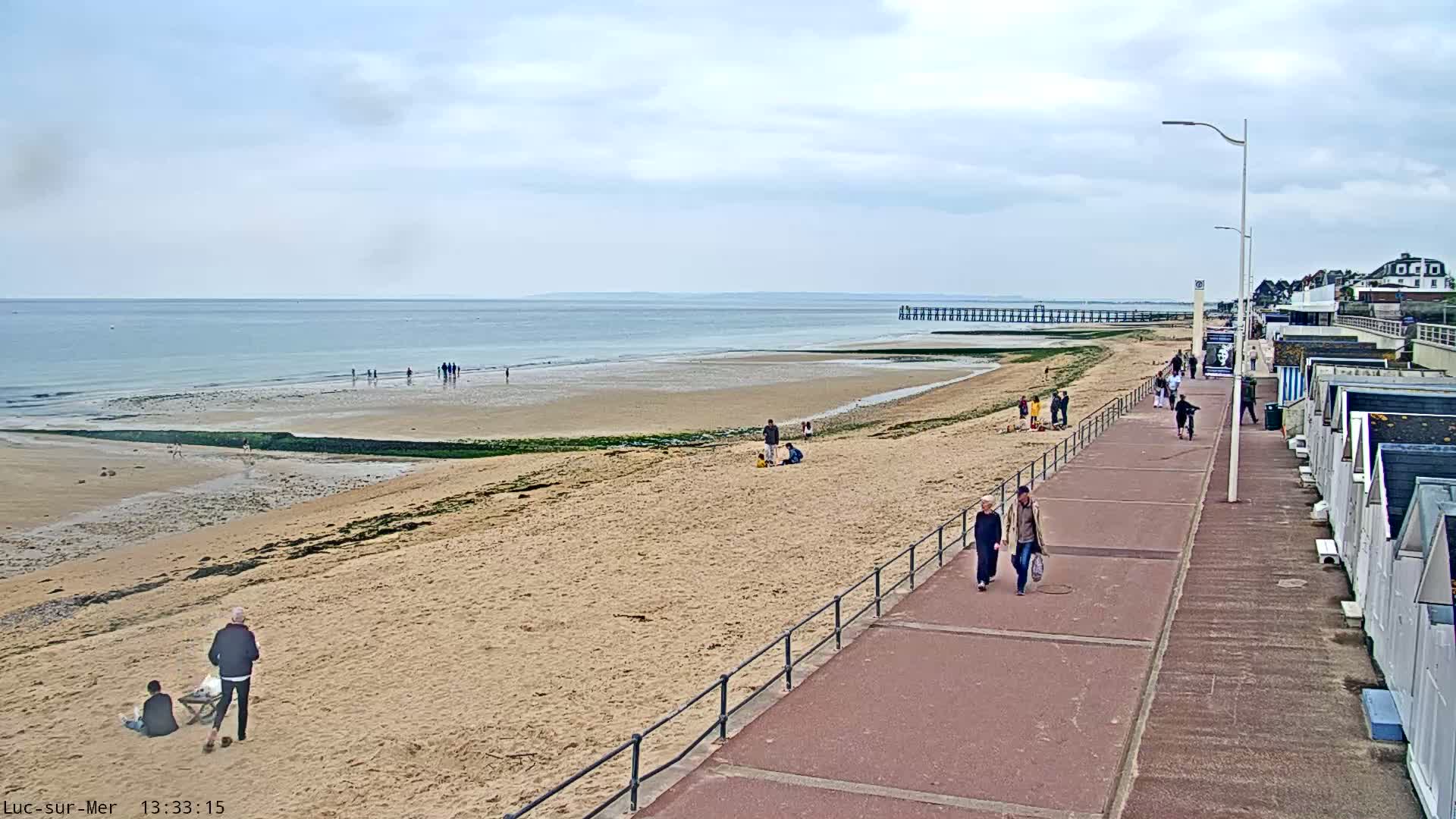 A sandy beach with a few people on it and a paved walkway lined with beach huts stretches alongside a calm sea under a mostly cloudy sky.