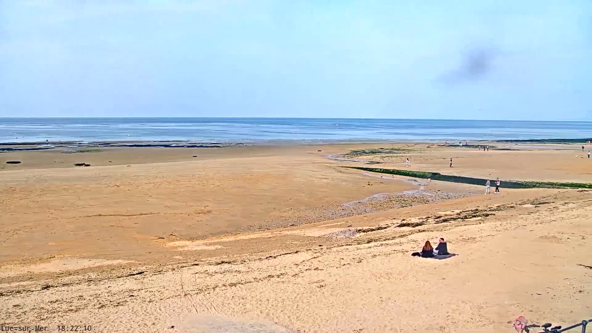 A wide sandy beach stretches toward a calm ocean under a mostly clear sky, with a few people scattered across the sand.