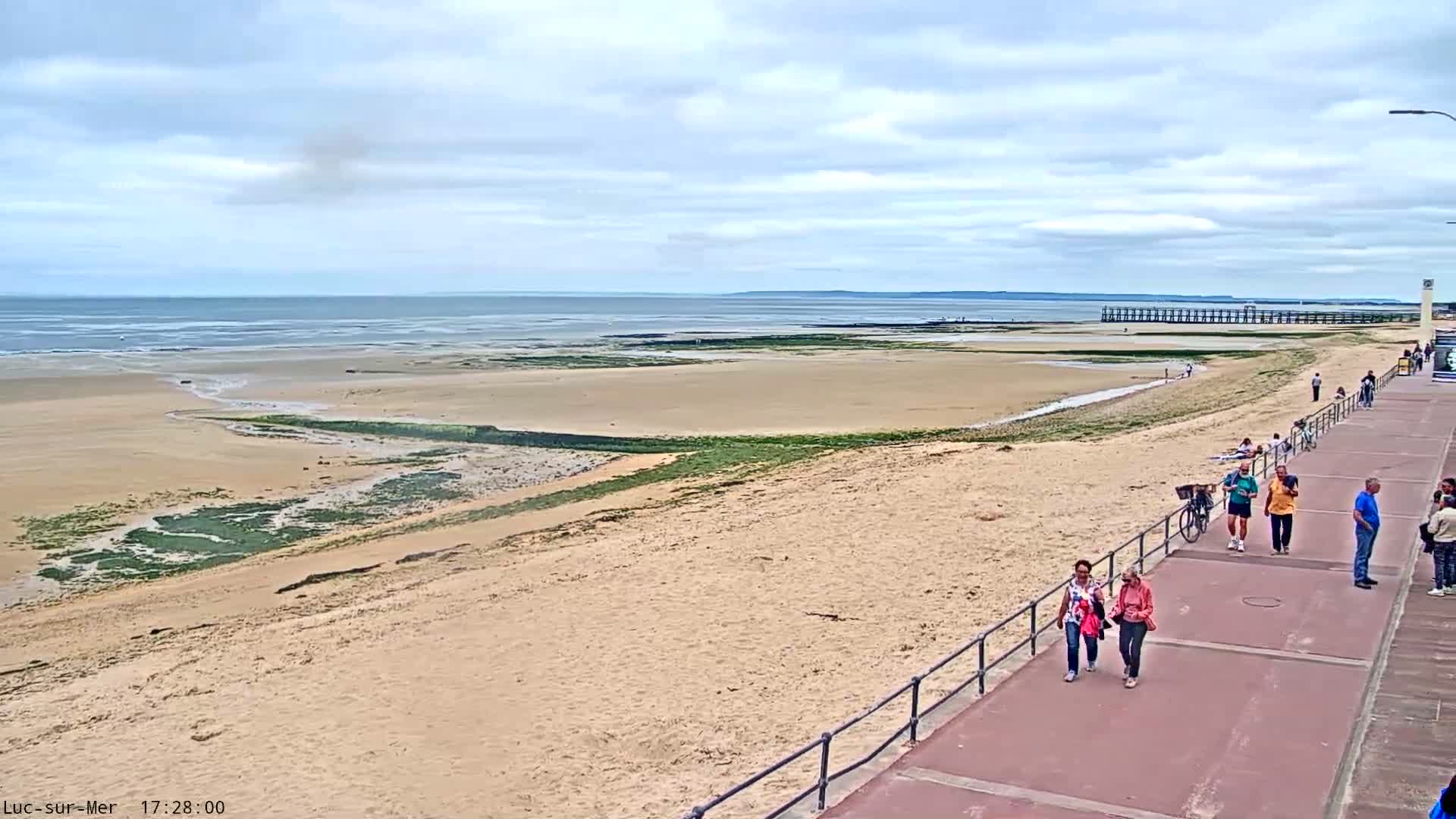 A sandy beach with a boardwalk and several people walking along it stretches to a calm ocean under a mostly cloudy sky.