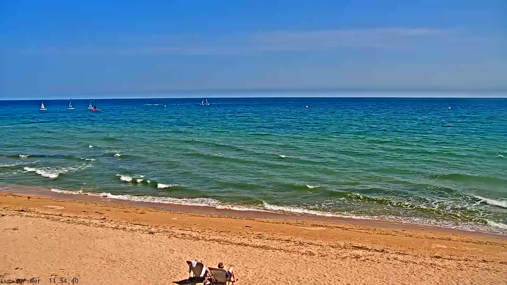 A sandy beach with two people sitting in chairs, overlooking a calm, teal-green ocean with several sailboats in the distance under a clear blue sky.