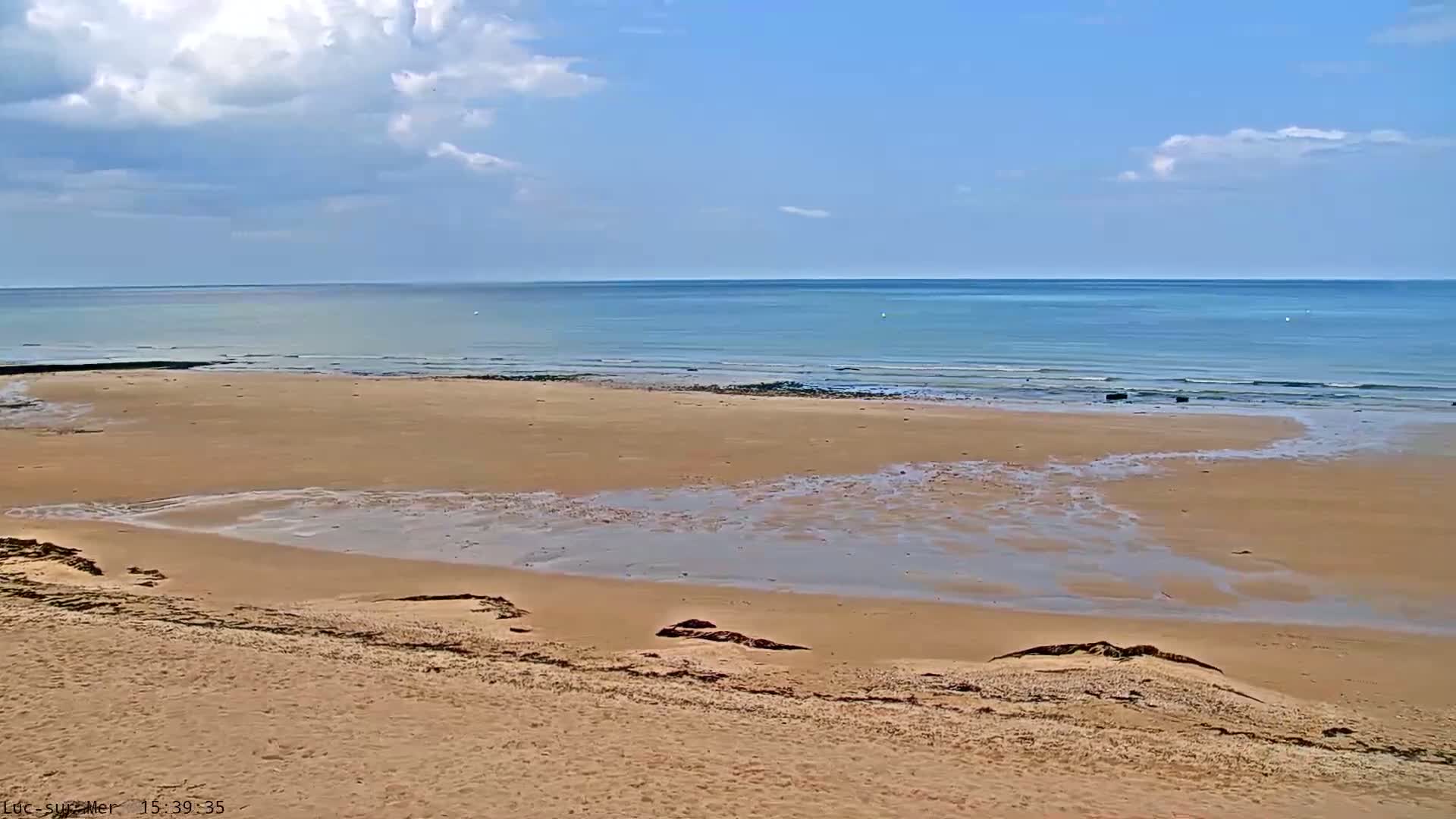 A mostly sunny day shows a calm ocean meeting a wide, sandy beach with small pools of water left by the receding tide.