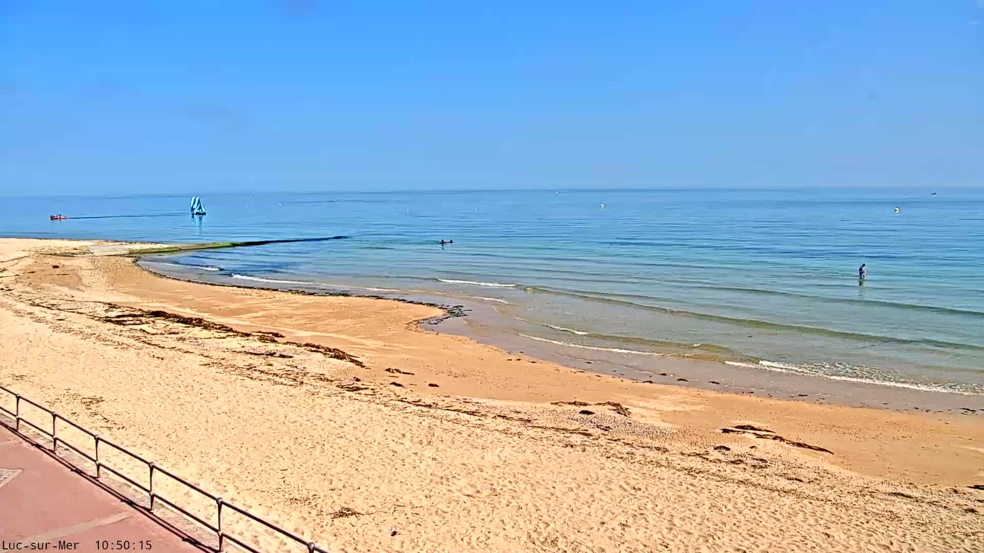 A sandy beach slopes down to a calm, light blue ocean on a sunny day, with a few people in the water and a small sailboat visible in the distance.