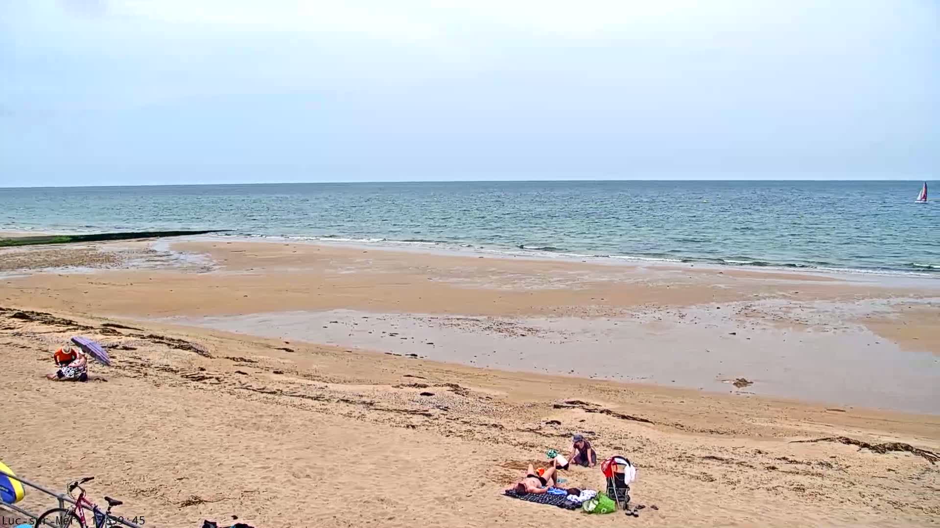A sandy beach stretches to a calm, blue ocean under a mostly clear sky, with a few people relaxing on the sand.
