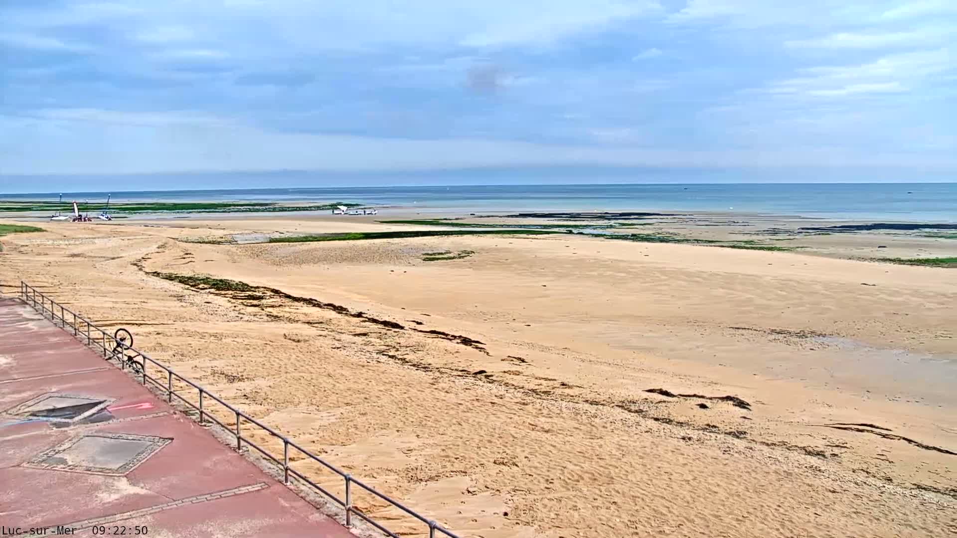 A sandy beach stretches to a calm ocean under a mostly cloudy sky, with a few small boats visible in the distance and a bicycle leaning against a railing nearby.