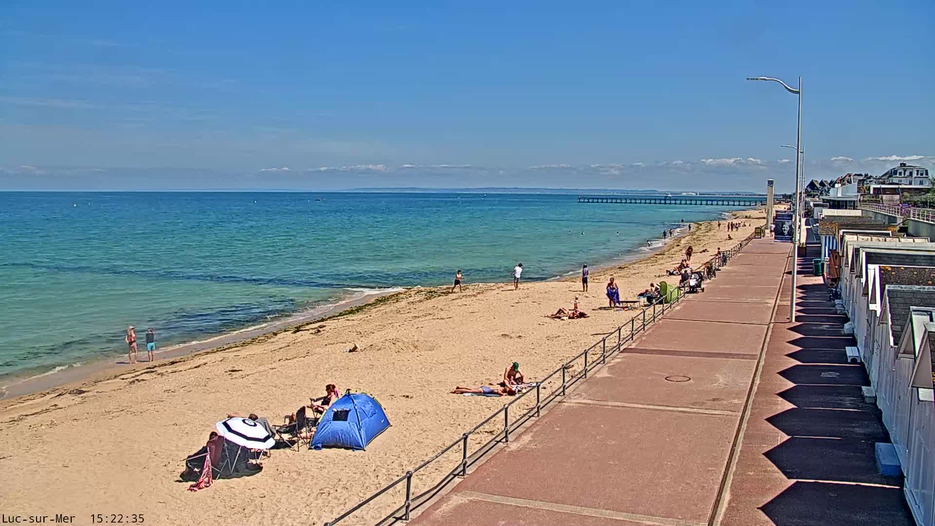 A sandy beach with people sunbathing and relaxing, a boardwalk with beach huts, and a calm ocean under a clear blue sky.