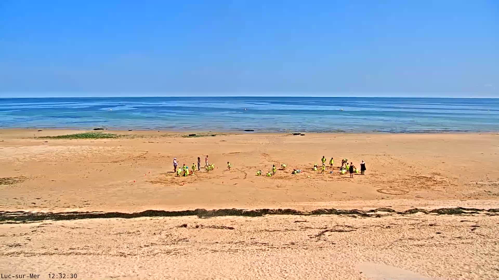 A group of children and adults are on a sandy beach near a calm, blue ocean under a clear, sunny sky.