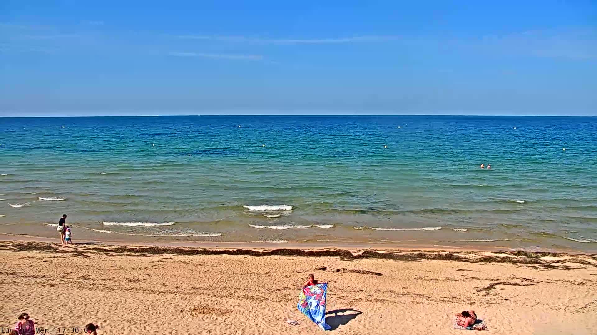 A sandy beach with a few people on it slopes down to a calm, teal ocean under a clear blue sky.