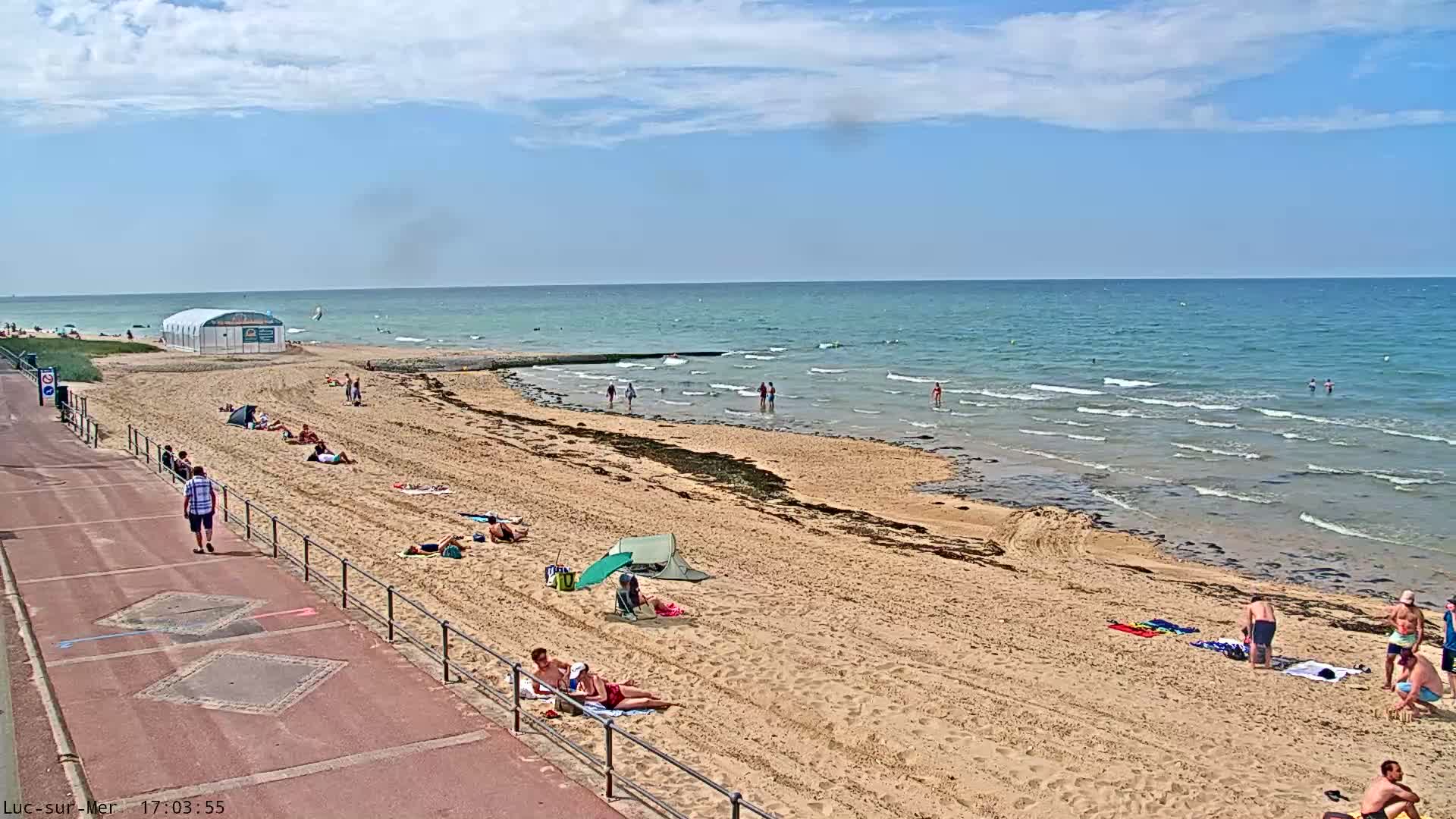 A sandy beach with people sunbathing and swimming in a calm ocean under a partly cloudy sky.