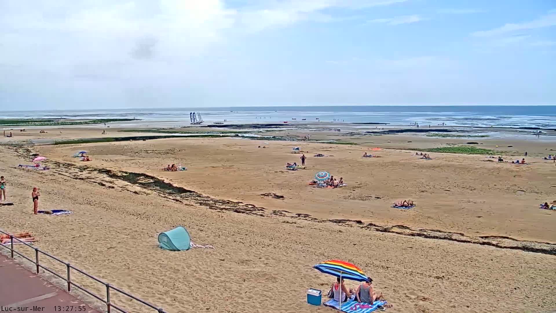 A sandy beach with scattered sunbathers and a few sailboats visible in the distance on a partly cloudy day.