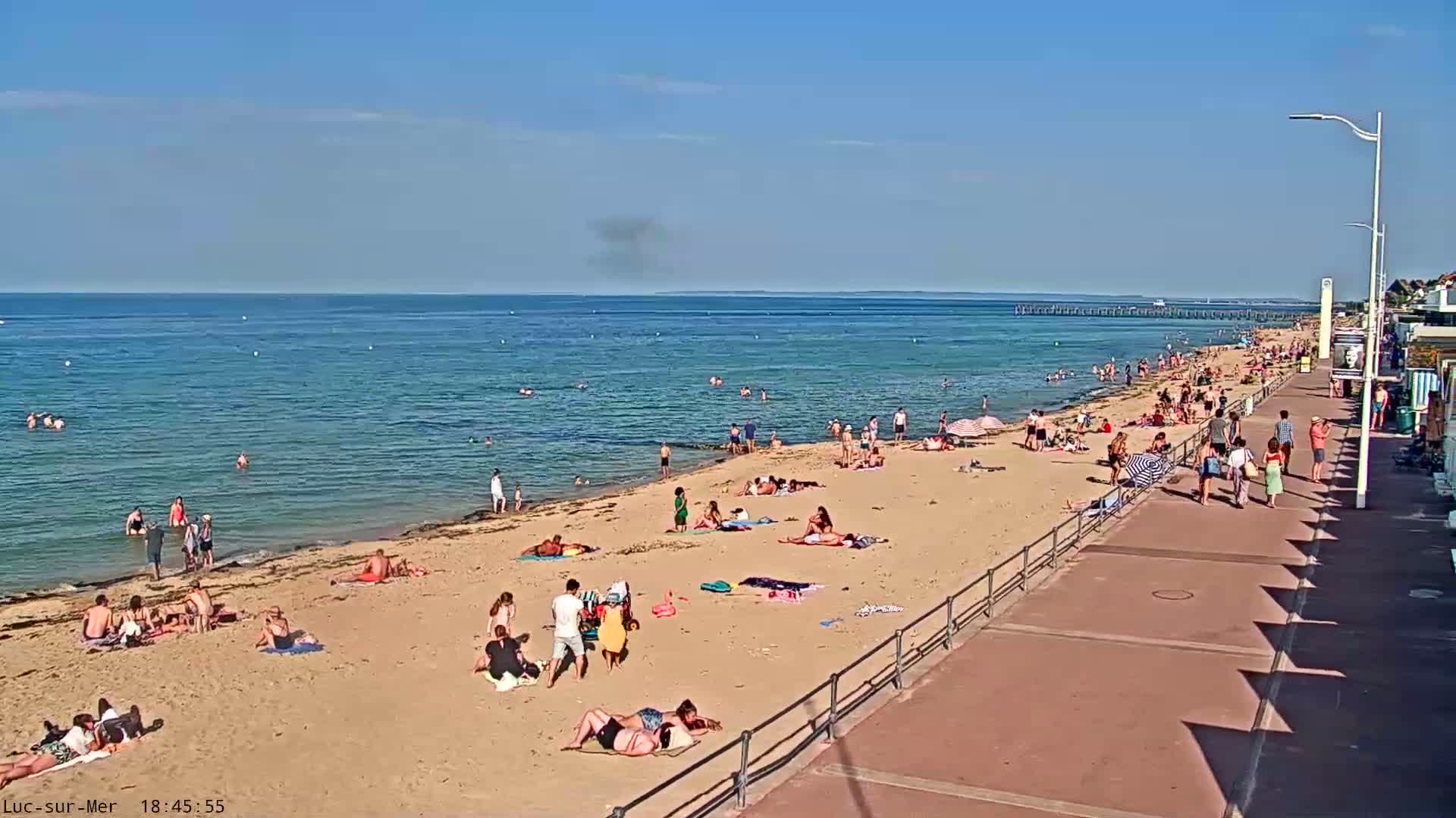 A sandy beach with many people sunbathing and swimming in a calm, clear ocean under a bright sunny sky.