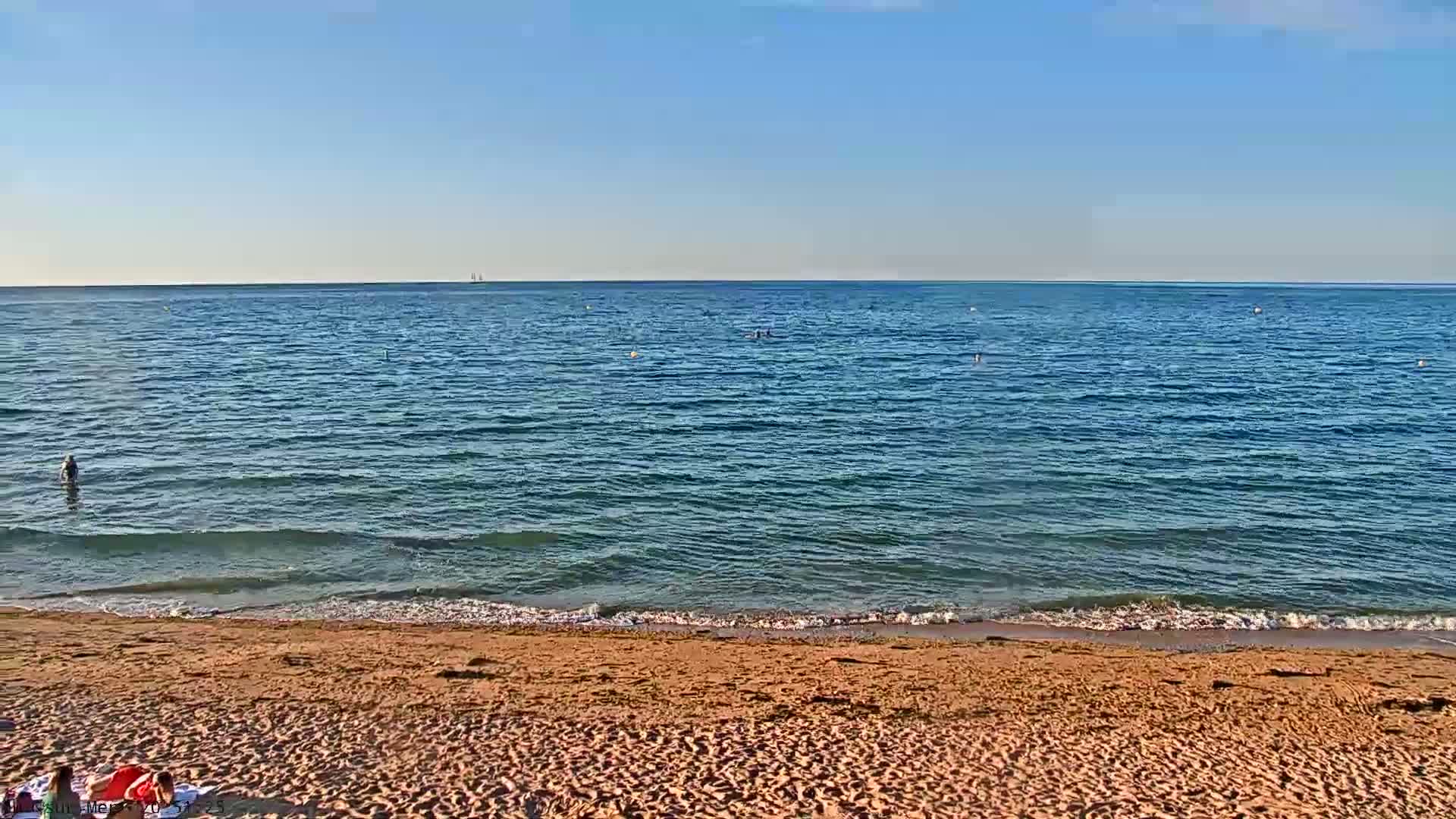 A sandy beach with calm, blue water and a few people swimming or standing in the ocean under a clear sky.