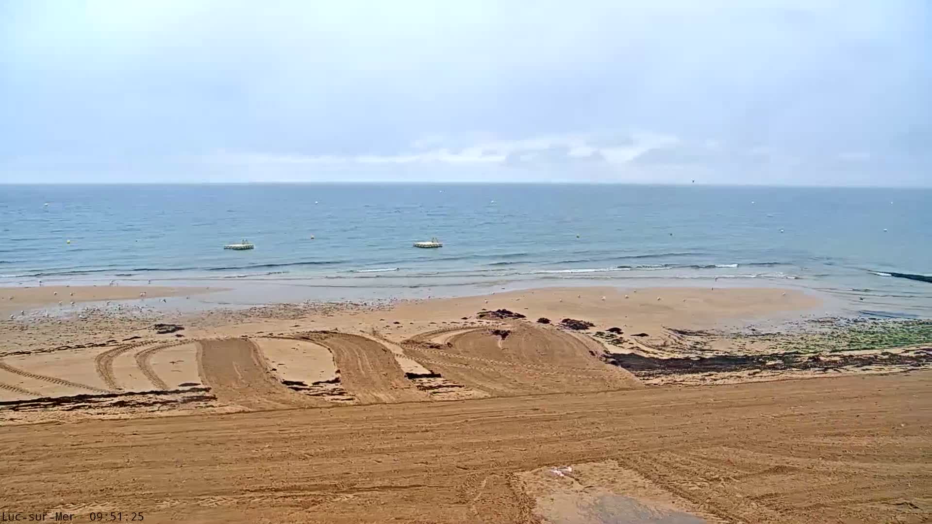 Tire tracks crisscross a sandy beach sloping down to a calm, light-blue ocean under a mostly cloudy sky.