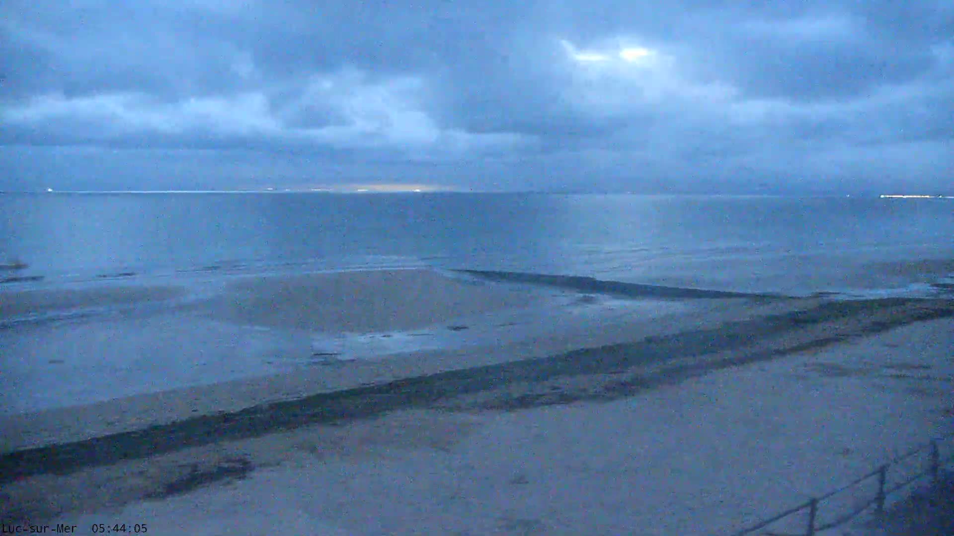 Tire tracks crisscross a sandy beach sloping down to a calm, light-blue ocean under a mostly cloudy sky.